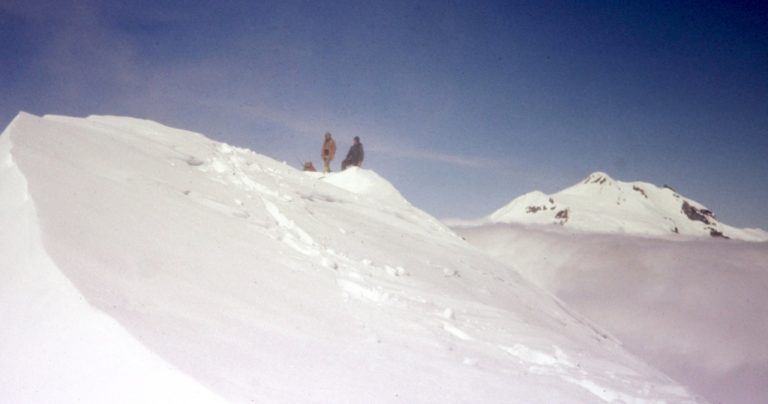 Two mountain climbers stand on the snowy summit of Chalangin Peak in the DaKobed-White Range