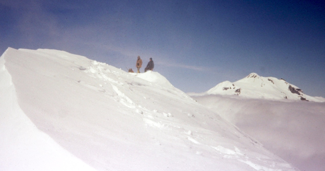 Two mountain climbers stand on the snowy summit of Chalangin Peak in the DaKobed-White Range