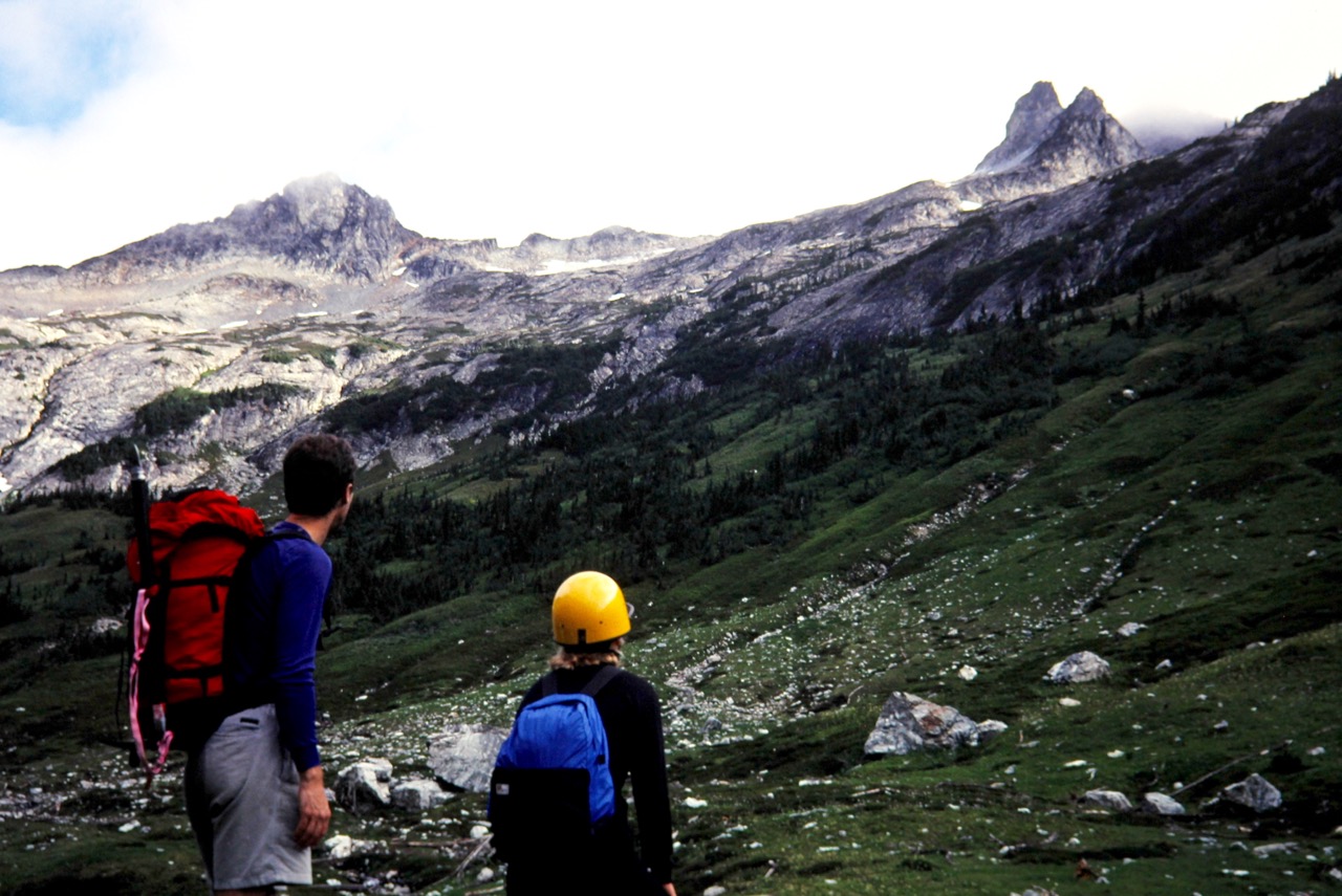 Two hikers look up at Dumbell Mountain from a large green meadow