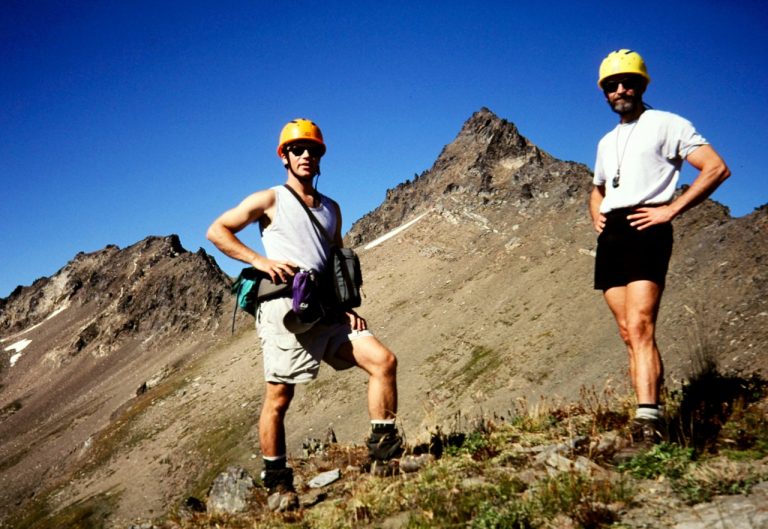 Two mountain climbers pose on a ridge with a rocky peak behind them