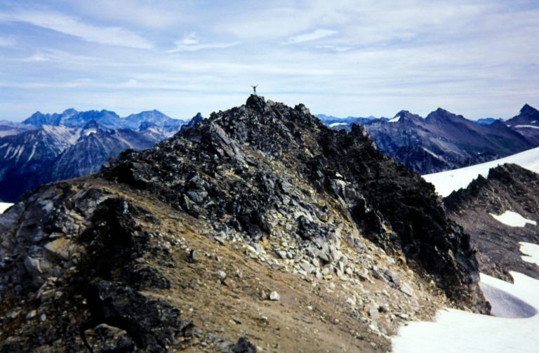 A lone mountain climber stands atop the rocky summit of Kololo Peak in the Glacier Peak Wilderness