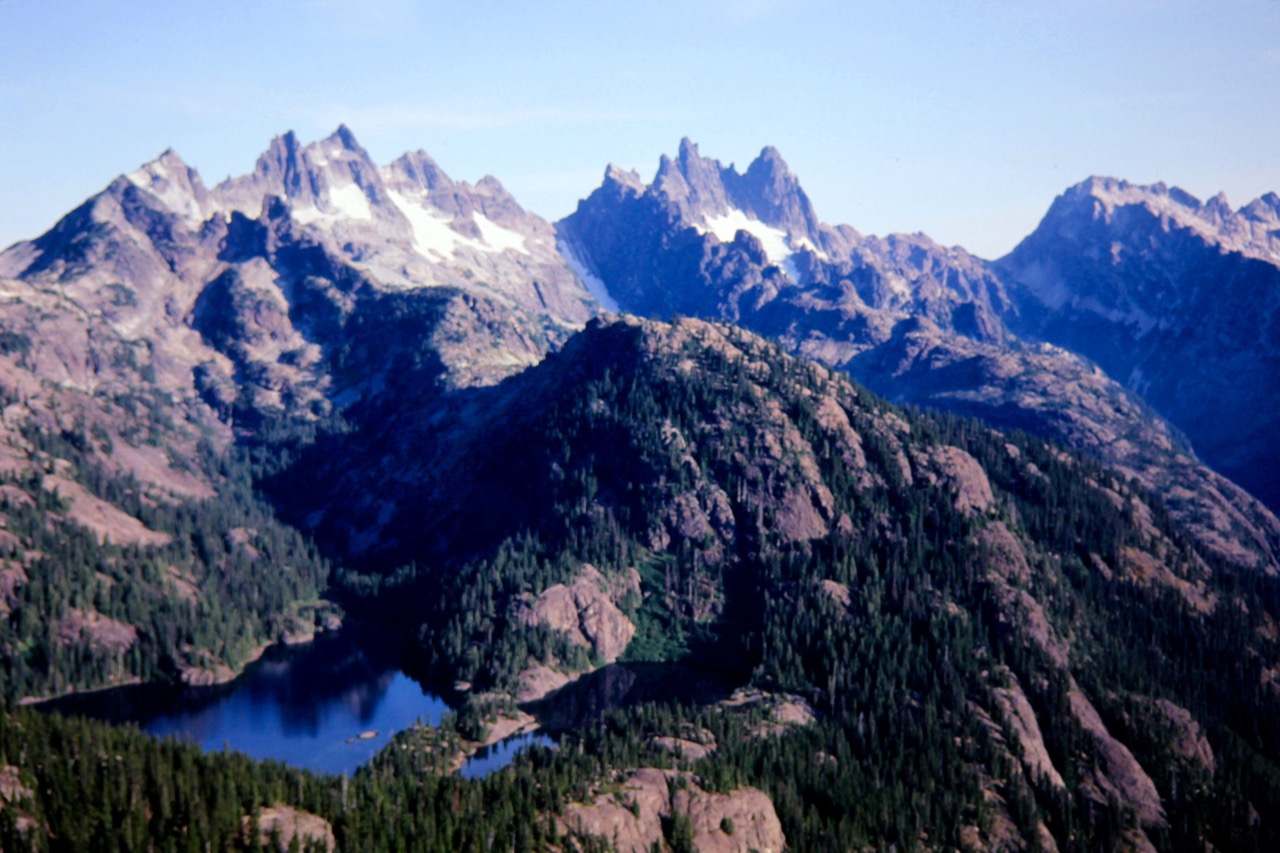 A row of the jagged Snoqualmie Mountains stand on the horizon with a blue lake below as seen from Three Queens Mountain