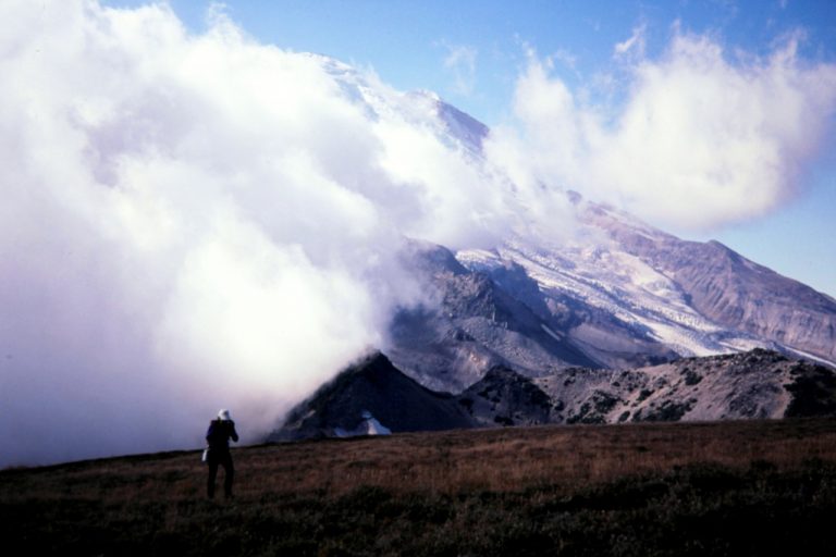 Clouds swarm around Mt Rainier as a hiker ascends a grassy slope on the way to Middle Cowlitz Chimney