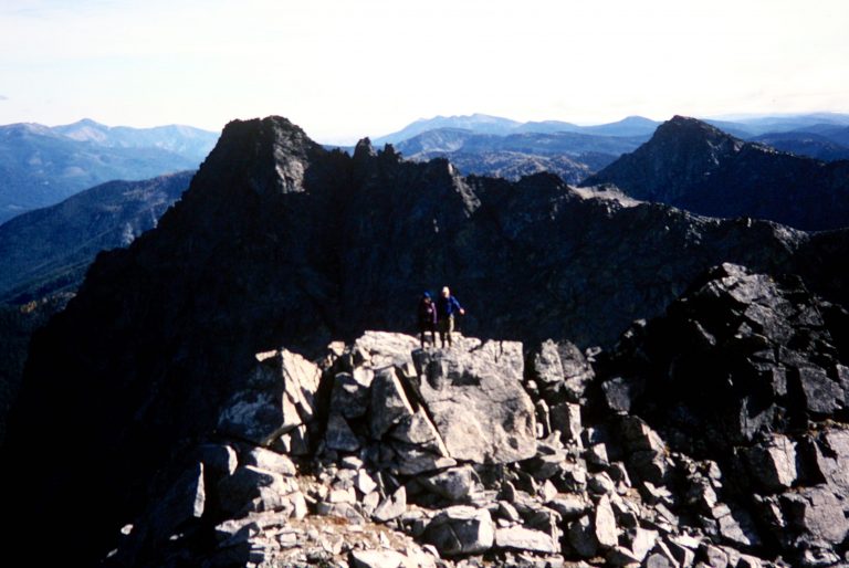 Mountain climbers stand on Rampart Mtn summit with a high peak behind them