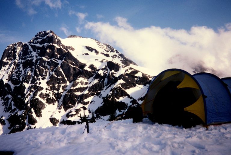 mountain climber's tent sits on a snowy ridge with Colonial Peak in North Cascades National Park behind