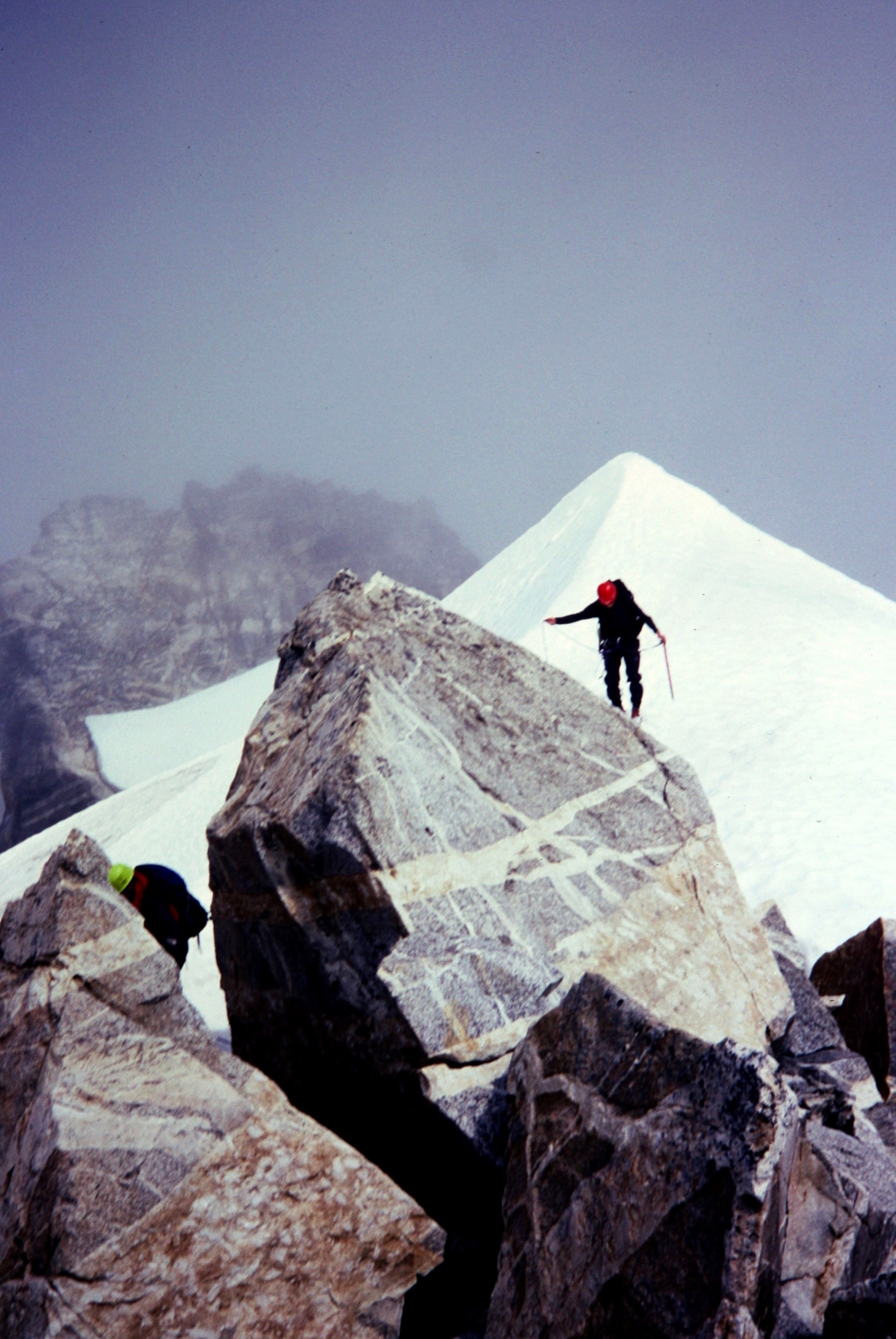 mountain climbers scrambling on the large summit boulders that make up East Mt Fury summit with snow fields in the background