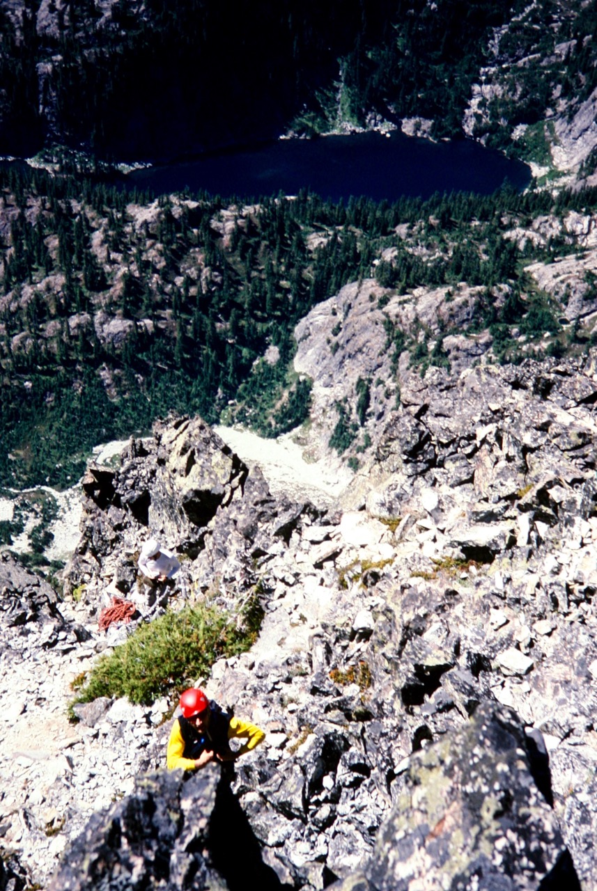 looking down on mountain climber completing the crux chimney on the rocky summit block of Bears Breast Mountain in the Alpine Lakes Wilderness with alpine lake and evergreen trees far below in the valley bottom