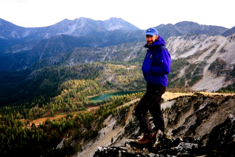 A blue clad climber stands on the summit of Martin Peak in the Sawtooth Mountains