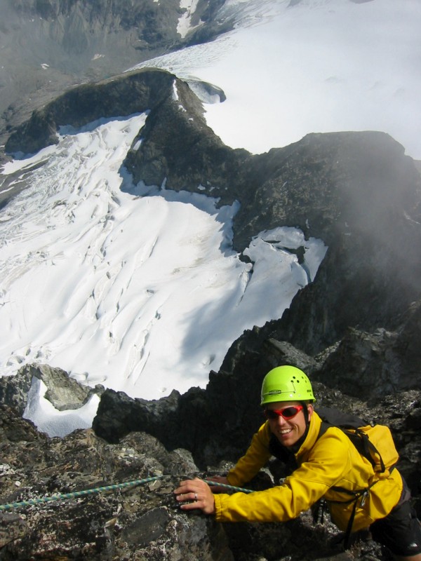 roped mountain climber making final moves to the Mt Redoubt summit above a steep rock face with snow slopes below