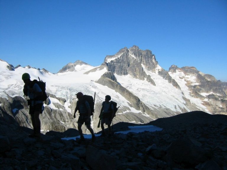 Three mountain climbers are silhouetted against Mt Redoubt as seen from Mt Spickard in the American Chilliwack Mountains