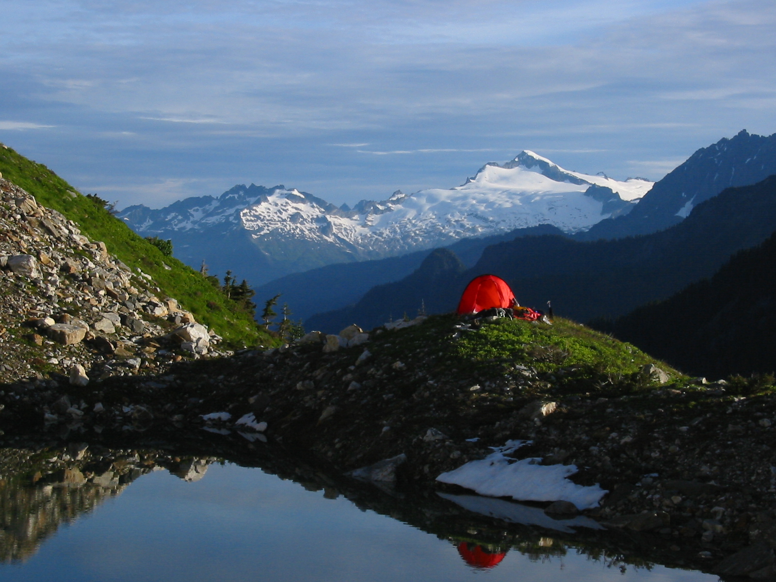 mountain climbers red tent on the grassy shoreline of a tarn camp with Eldorado Peak in the distance