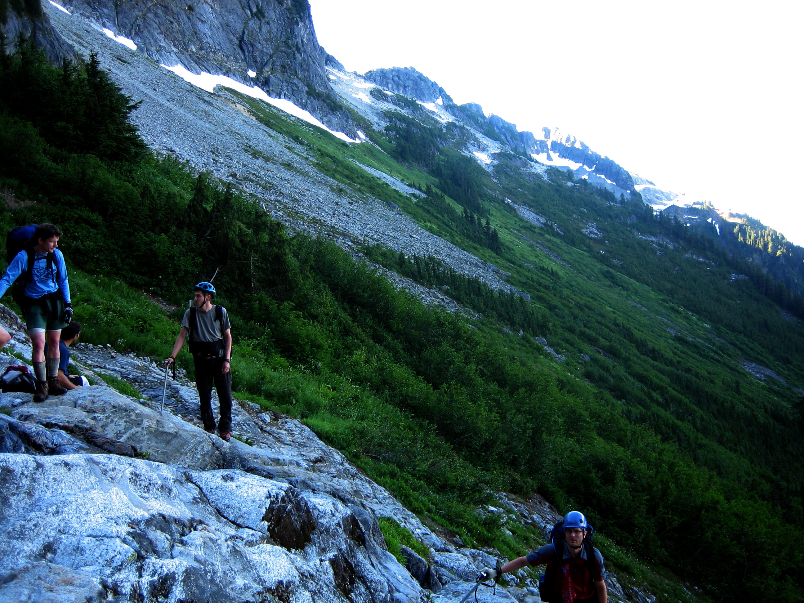 mountain climbers syanding on rock slabs with green bushes, scree fields, and small snow patches in the background in the Lower Skagit Mountains