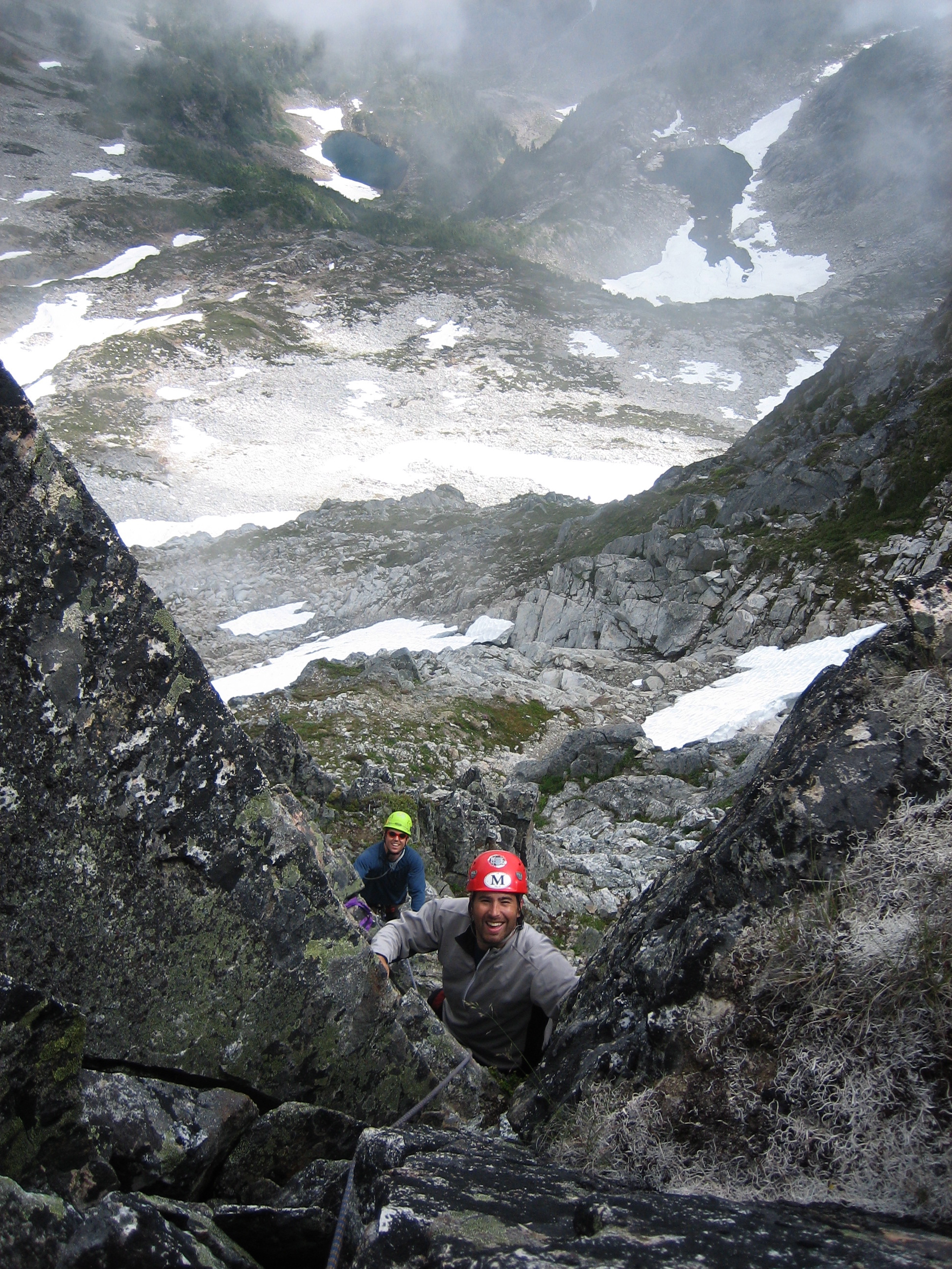looking down on roped mountain climbers ascending crack on summit block on Mt Despair in the Lower Skagit Mountains with steep rock, snow patches, and wafting clouds in North Cascades National Park
