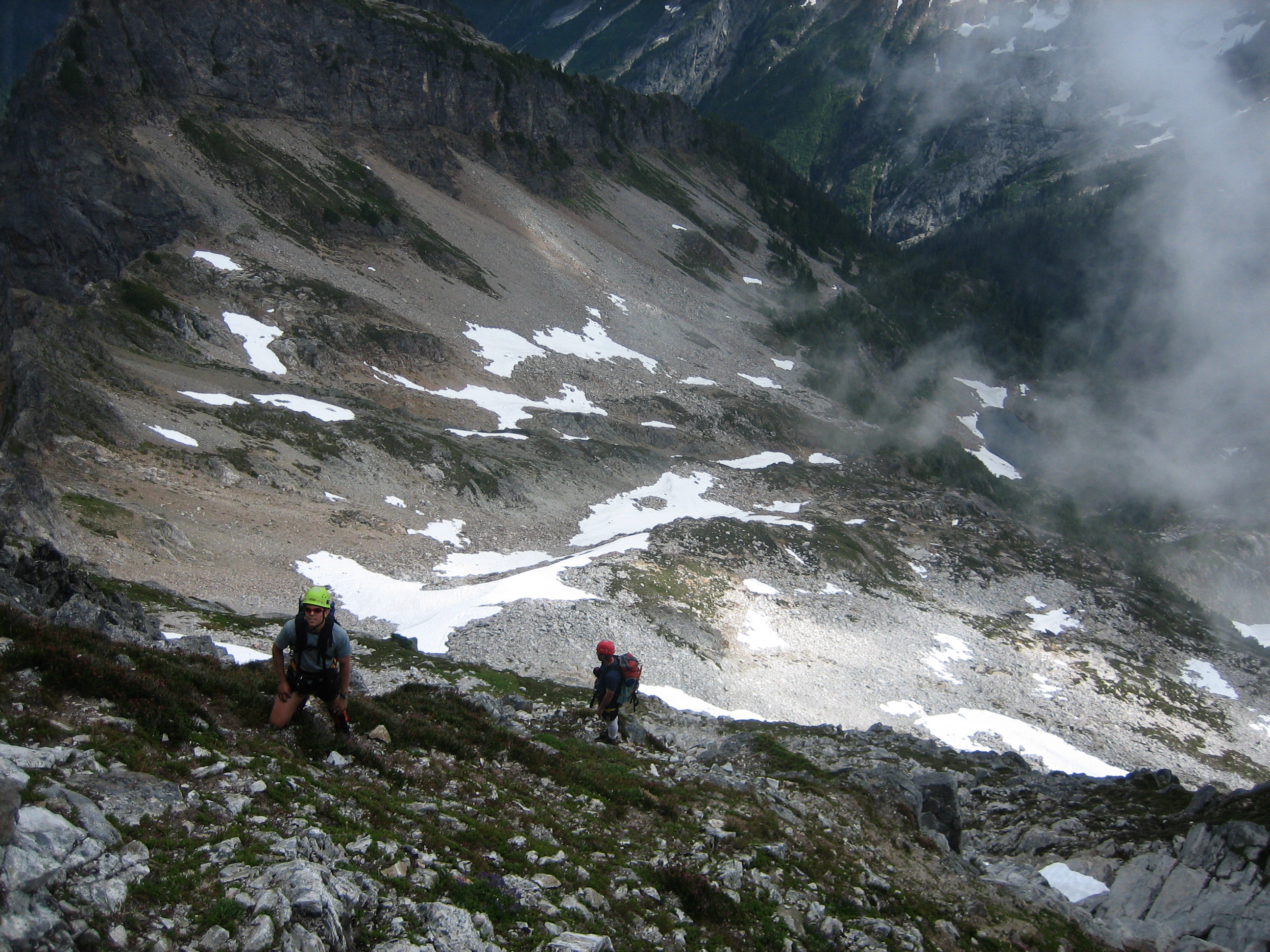 looking down on mountain climbers scrambling steep rock and heather on the face of Mt Despair in the Lower Skagit Mountains with snow and scree filled basin below in North Cascades National Park