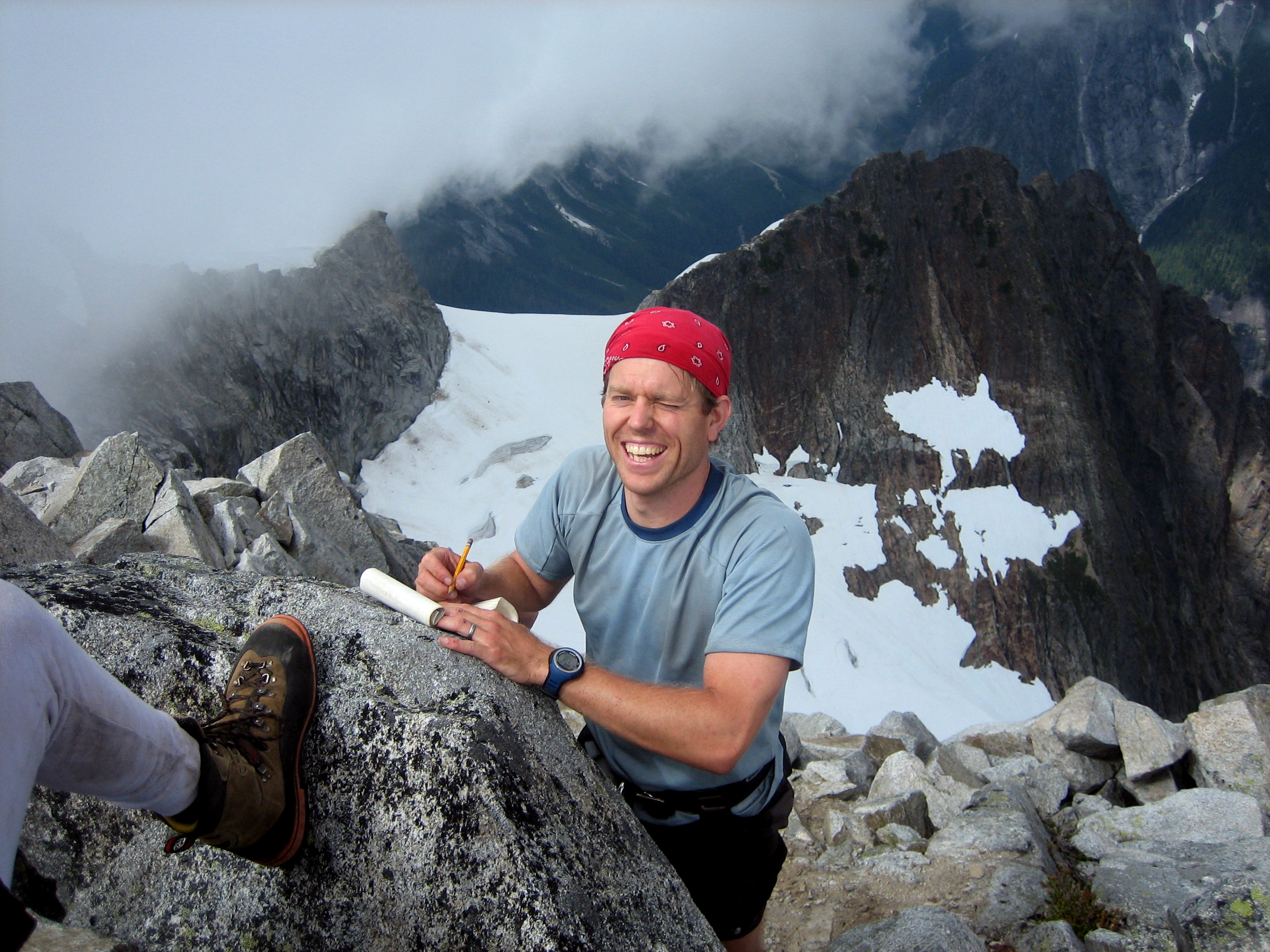mountain climber signing Mt Despair registar on the rock horn summit of Mt Despair in the Lower Skagit Mountains in North Cascades National Park with snow covered rock ridges and wafting clouds in the background 