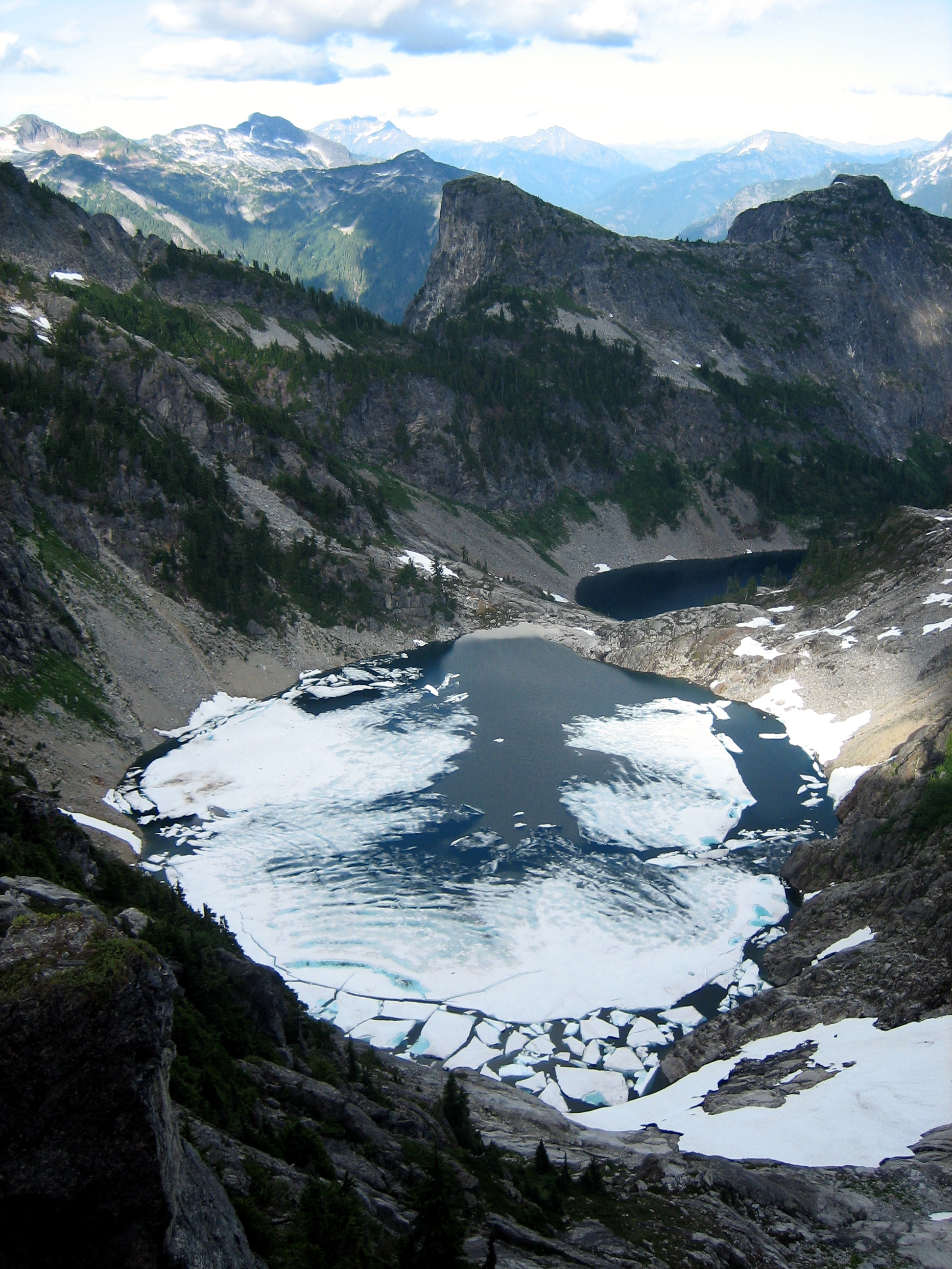 Looking down on partially melted out Thornton Lakes with steep rock walls and patches of heather as seen from mountain climber's camp on Thornton Ridge in the Lower Skagit Mountains in North Cascades National Park