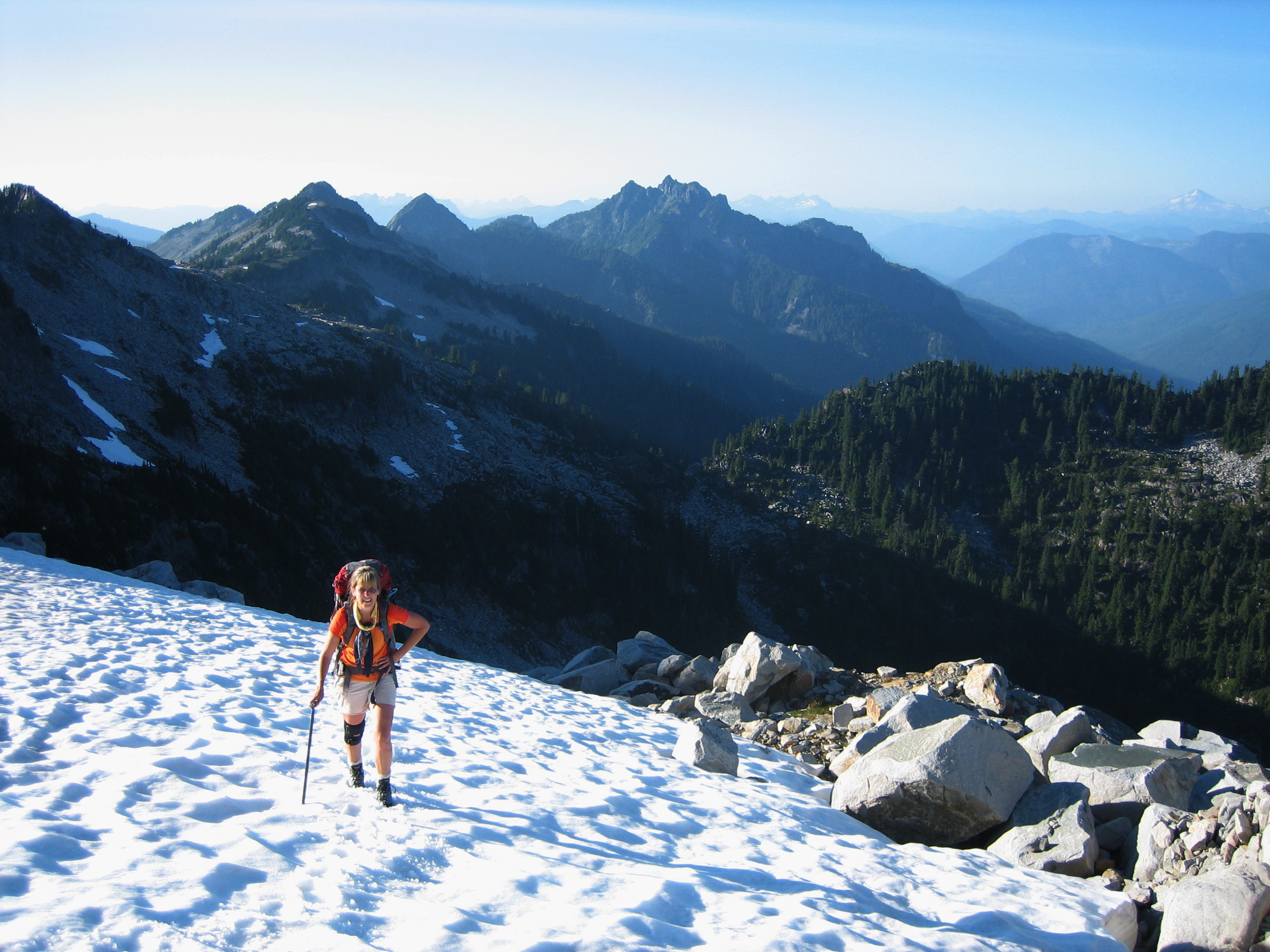 hiker botting up snow field toward Camp Robber Peak with boulder field and mountains in the background