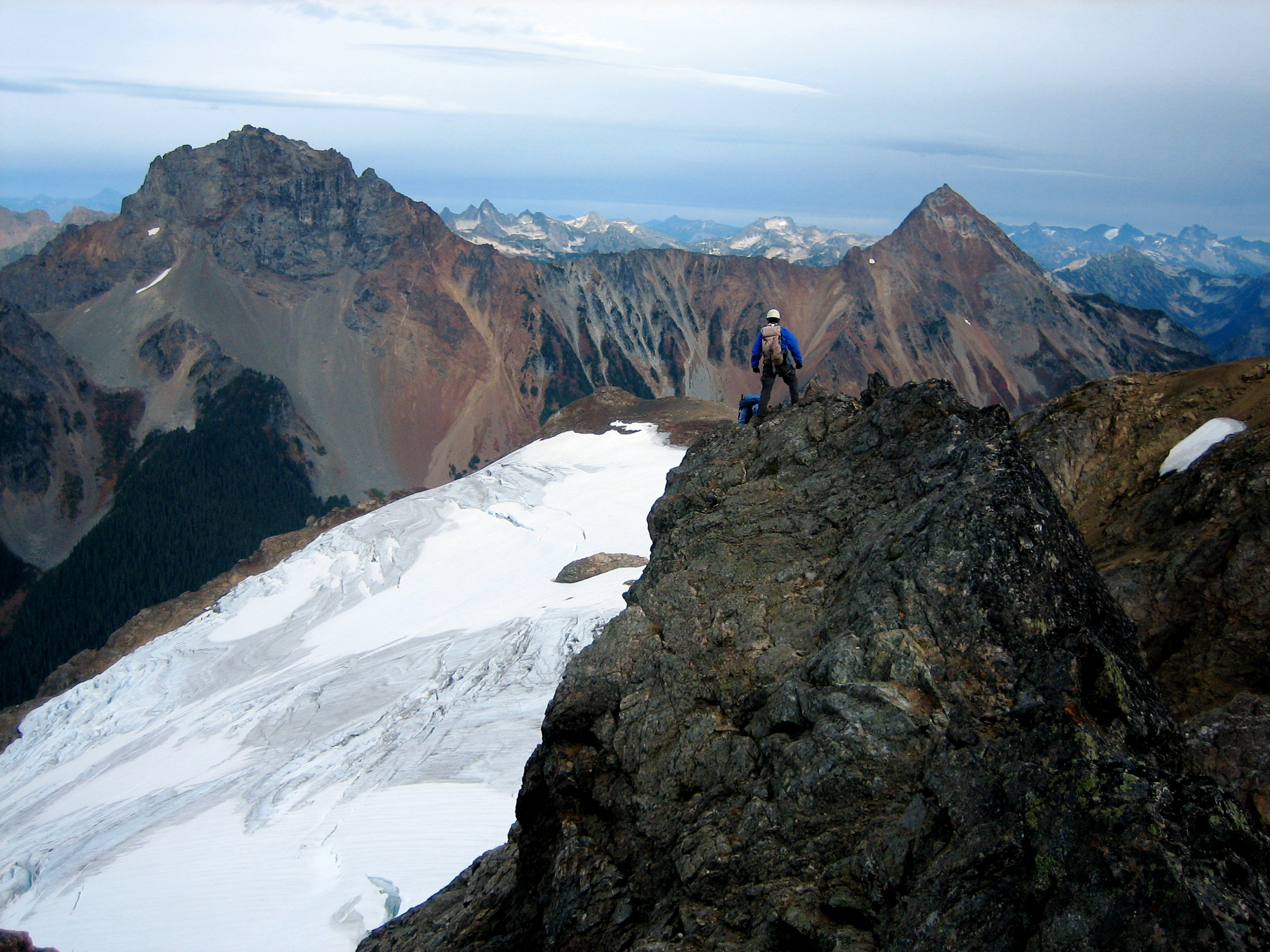 American Border Peak and Mt Larrabee in the background with a mountain climber scrambling the rock crest of Tomyhoi Peak