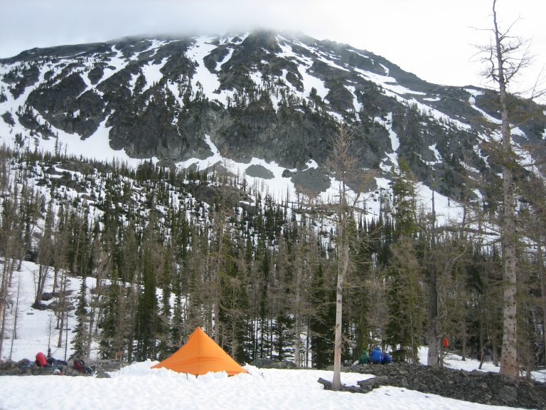 An orange tent sits in a snowy basin below the broad face of Big Craggy Peak