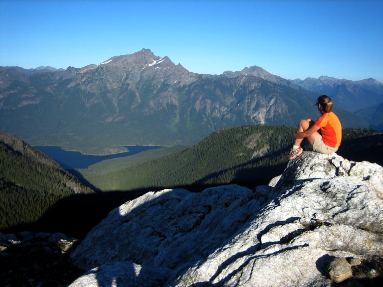 A woman hiker sits on a white rock and looks across Ross Lake at Jack Mountain