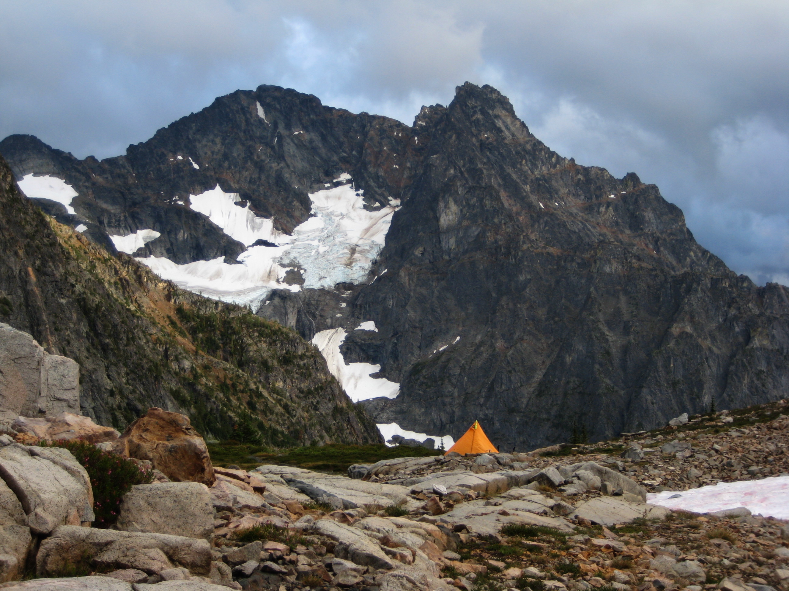 mountain climber's orange tent pitched on the slabby shoreline of Silent Lakes in North Cascades National Park with Black Peak in the Ragged RIdge mountain range filling the skyline