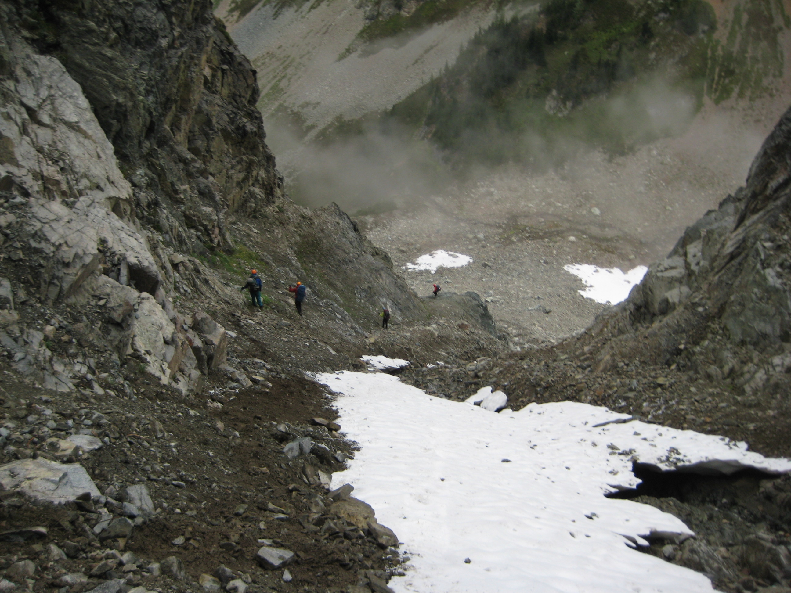 looking down on mountain climbers descending steep rock and dirt gulley leading out of Silent Lakes in North CAscades National Park with linguring snow fields and FIsher Basin below 
