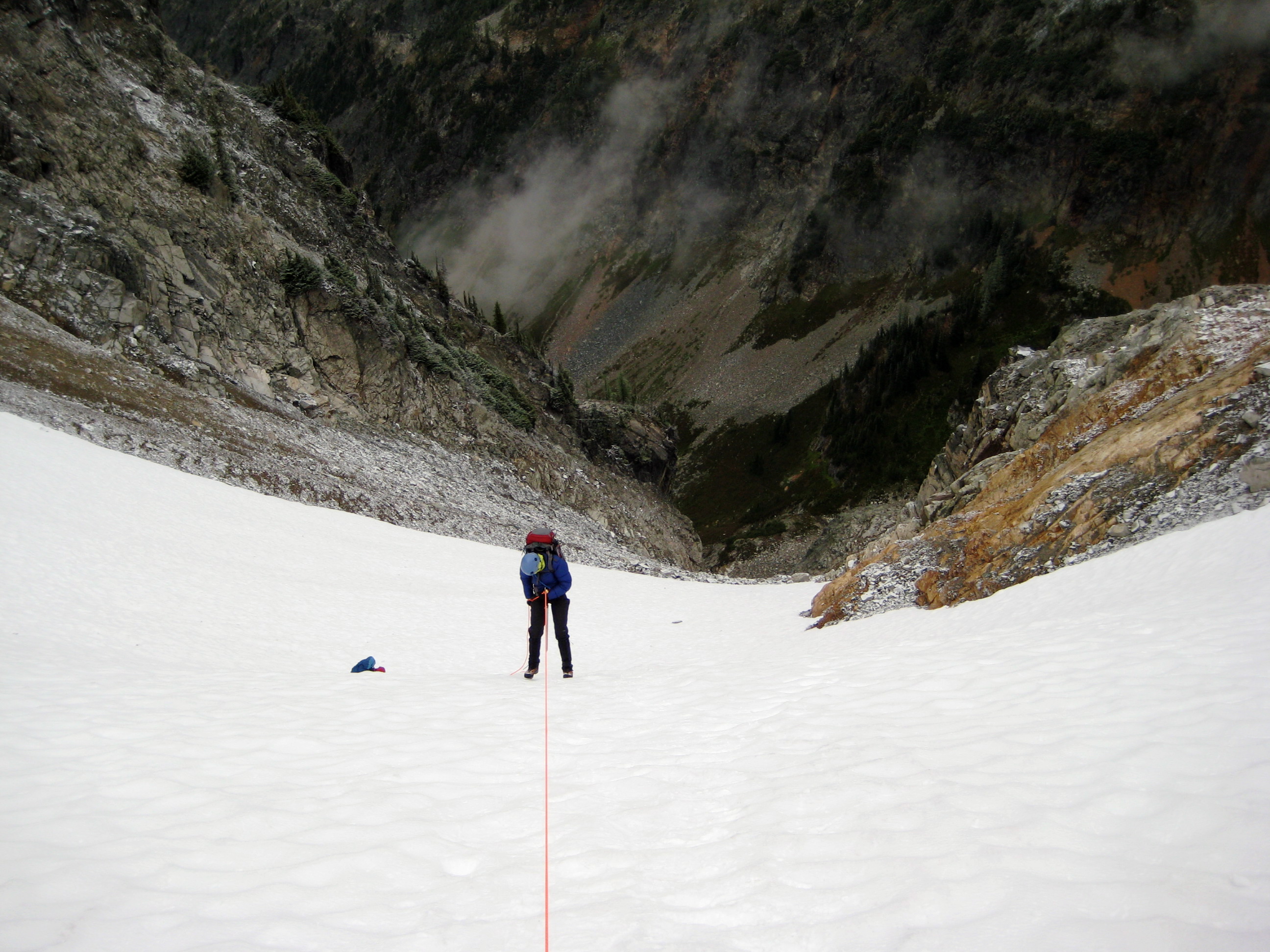 looking down on mountain climber rappelling steep snow slope with rock slabs on each side and the Fisher Basin below