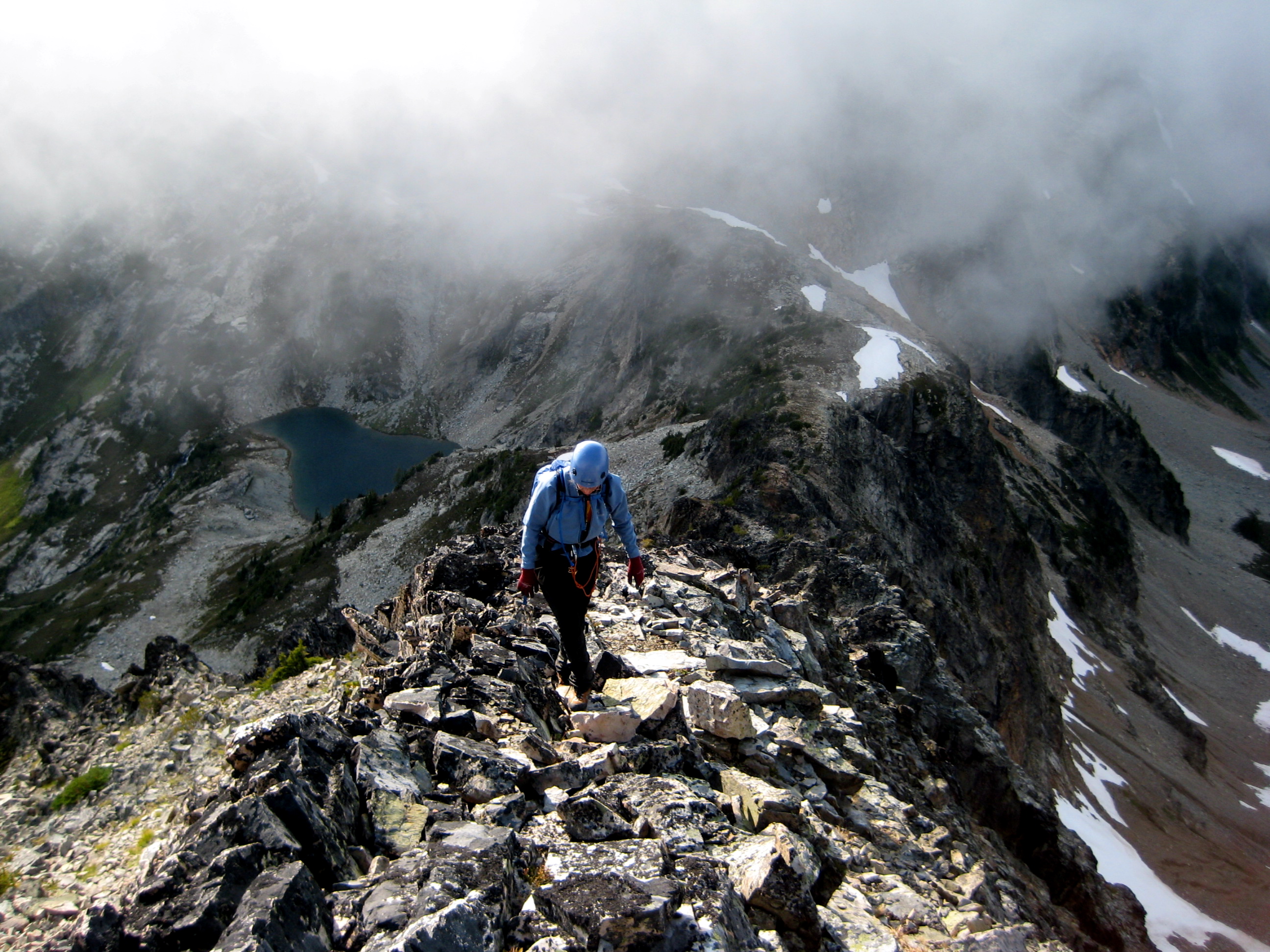 looking down on mountain climber scrambling Fisher Peak's broken rock ridge with Silent Lakes in North Cascades National Park being engulfed by the clouds