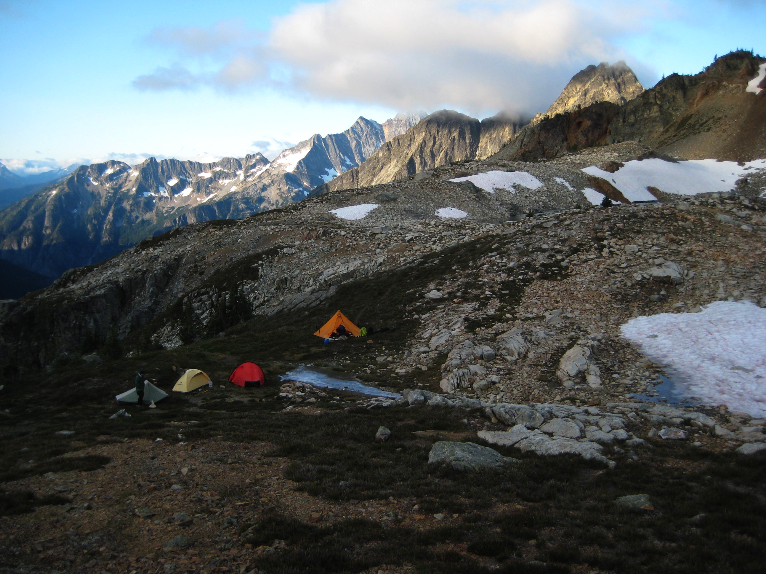 morning light on mountain climber's camp on the rocky shore of Silent Lakes in North Cascades National Park with linguring snow fields and the Ragged Ridge mountain range in the background