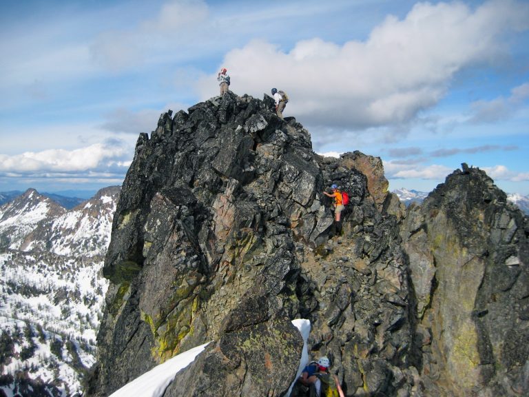 Several mountain climbers scramble up the rocky summit of Main Riddle Peak in Glacier Peak Wilderness on a blue sky day