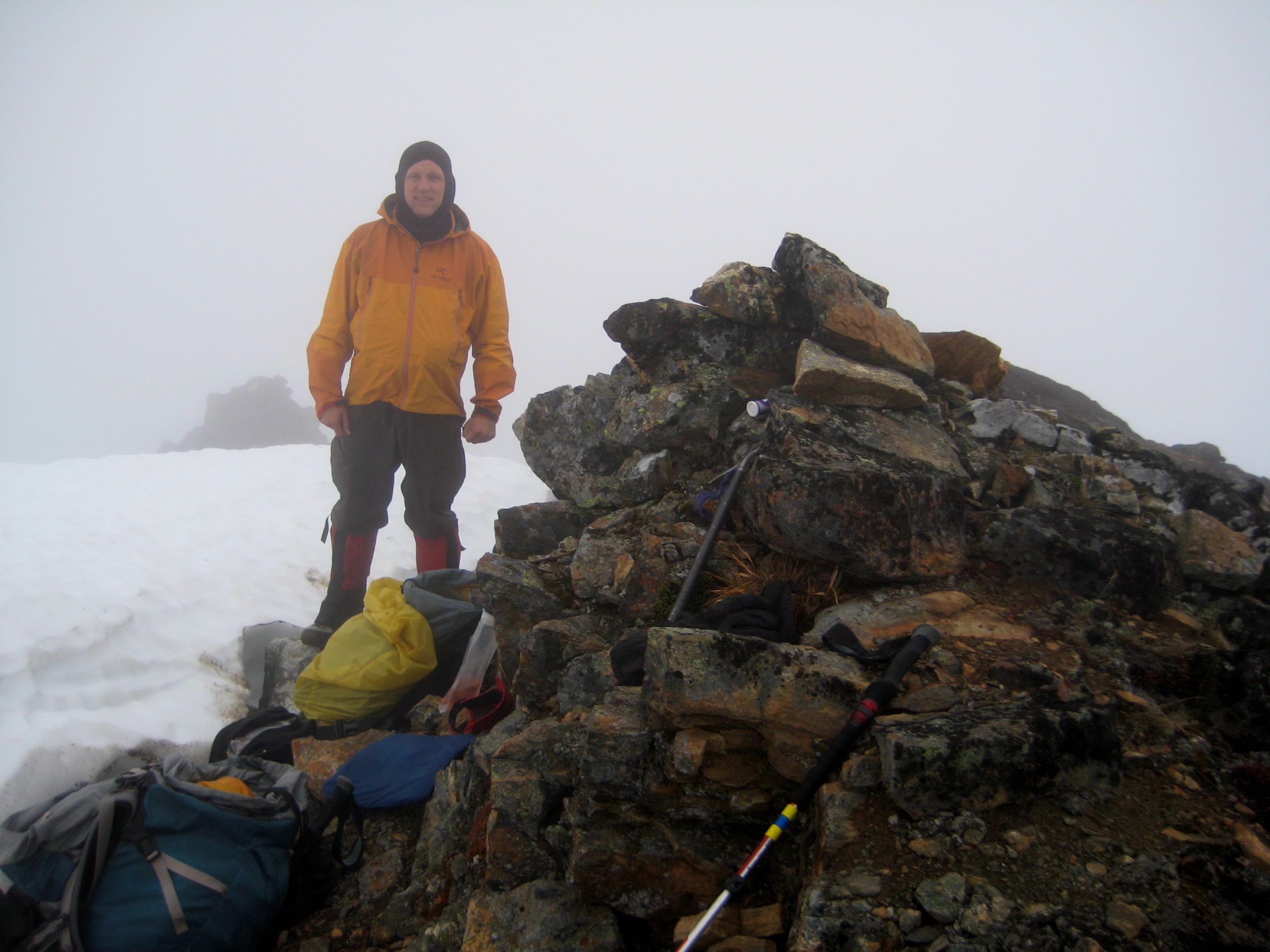 mountain climber in the fog stands next to the rocky summit of Mt Prophet in North Cascades National Park with snow fields in the background