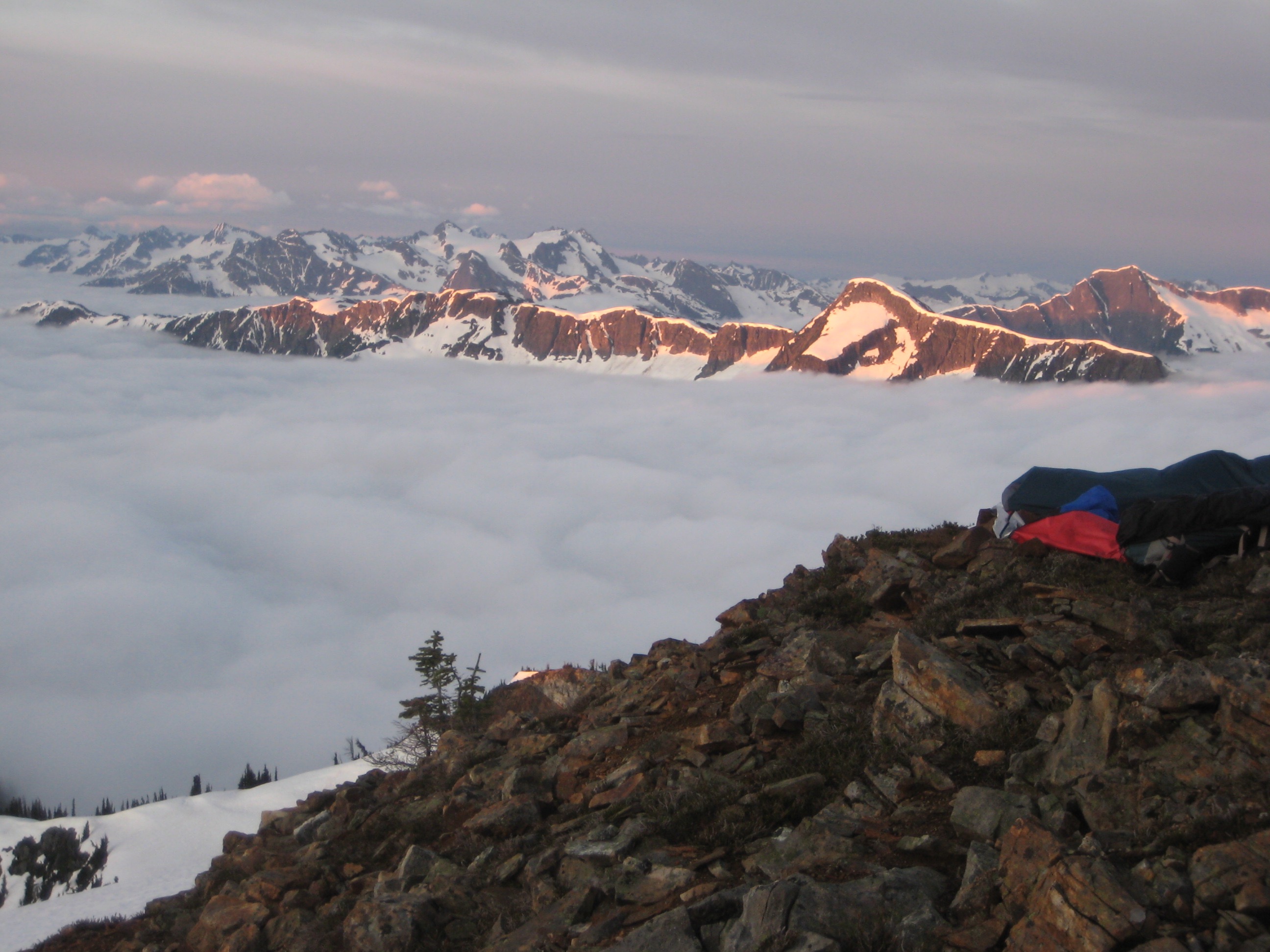 mountain climbers bivy camp on the rocky shoulder of Mt Prophet with snowy mountains of North Cascades National Park in the background above valley fog