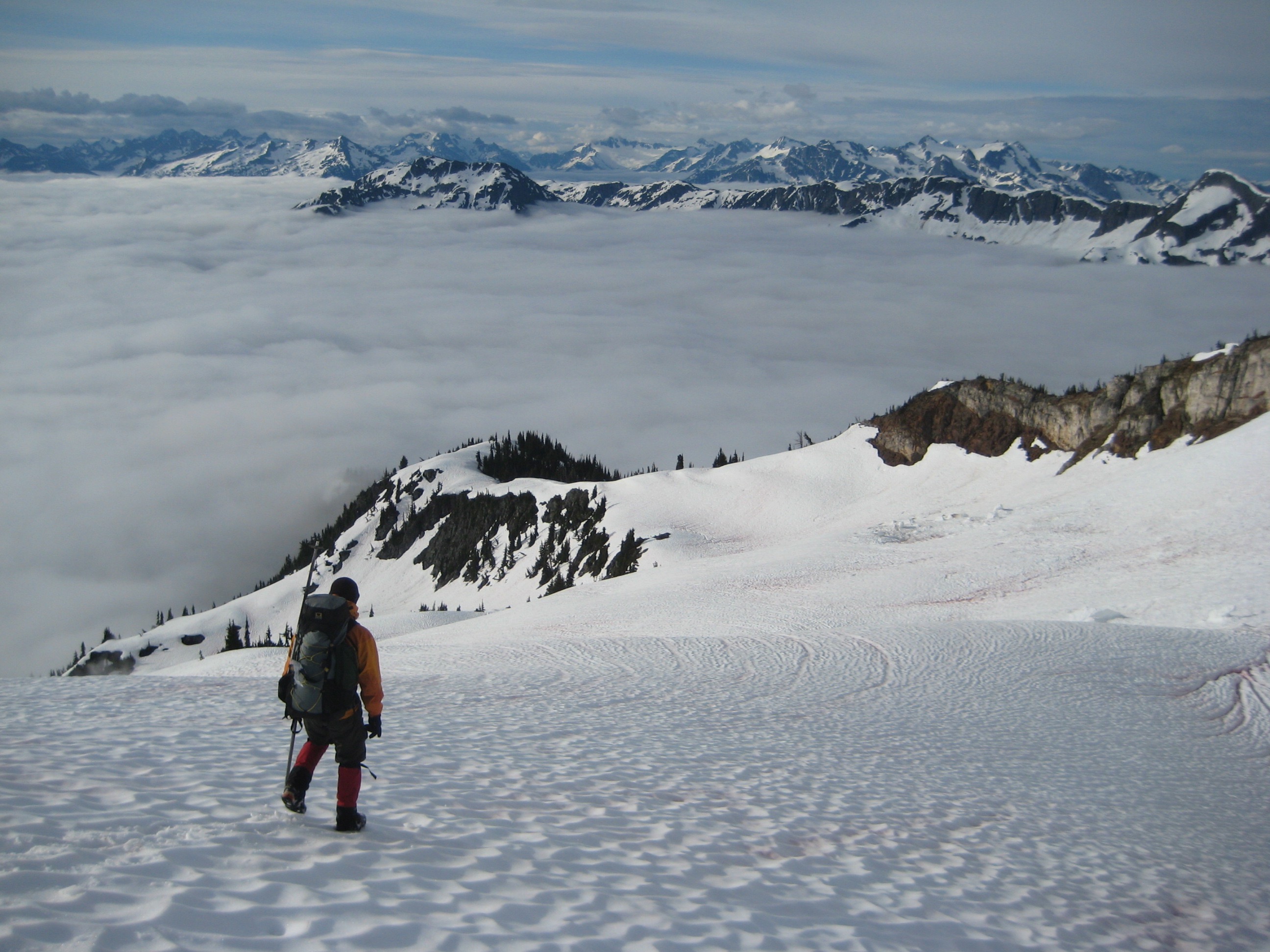mountain climber plunge stepping down snow field on Mt Prophet in North Cascades National Park with distant mountain views and valley fog
