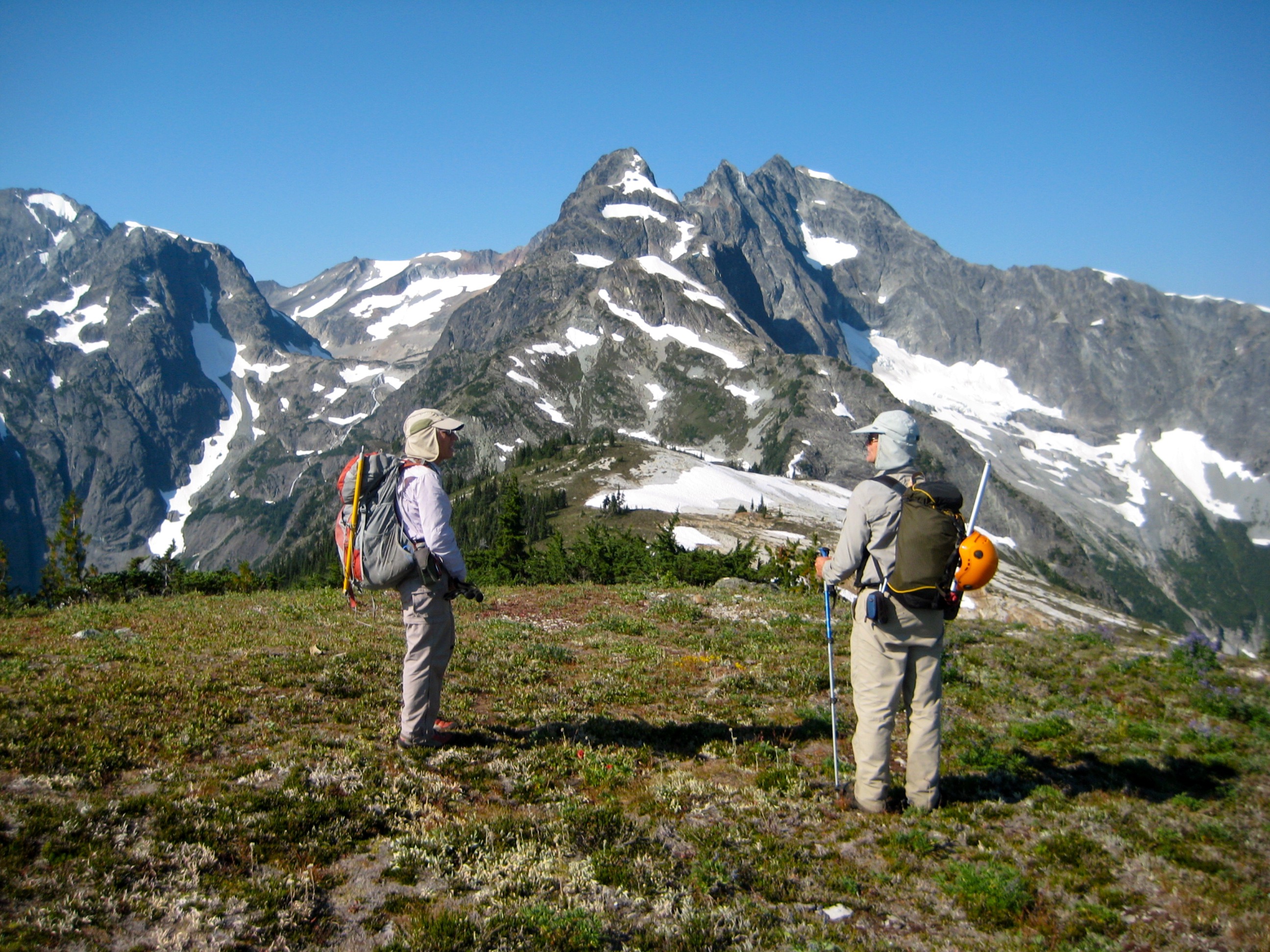 Two hikers stand on a grassy hill below the rugged Devils Tongue peak