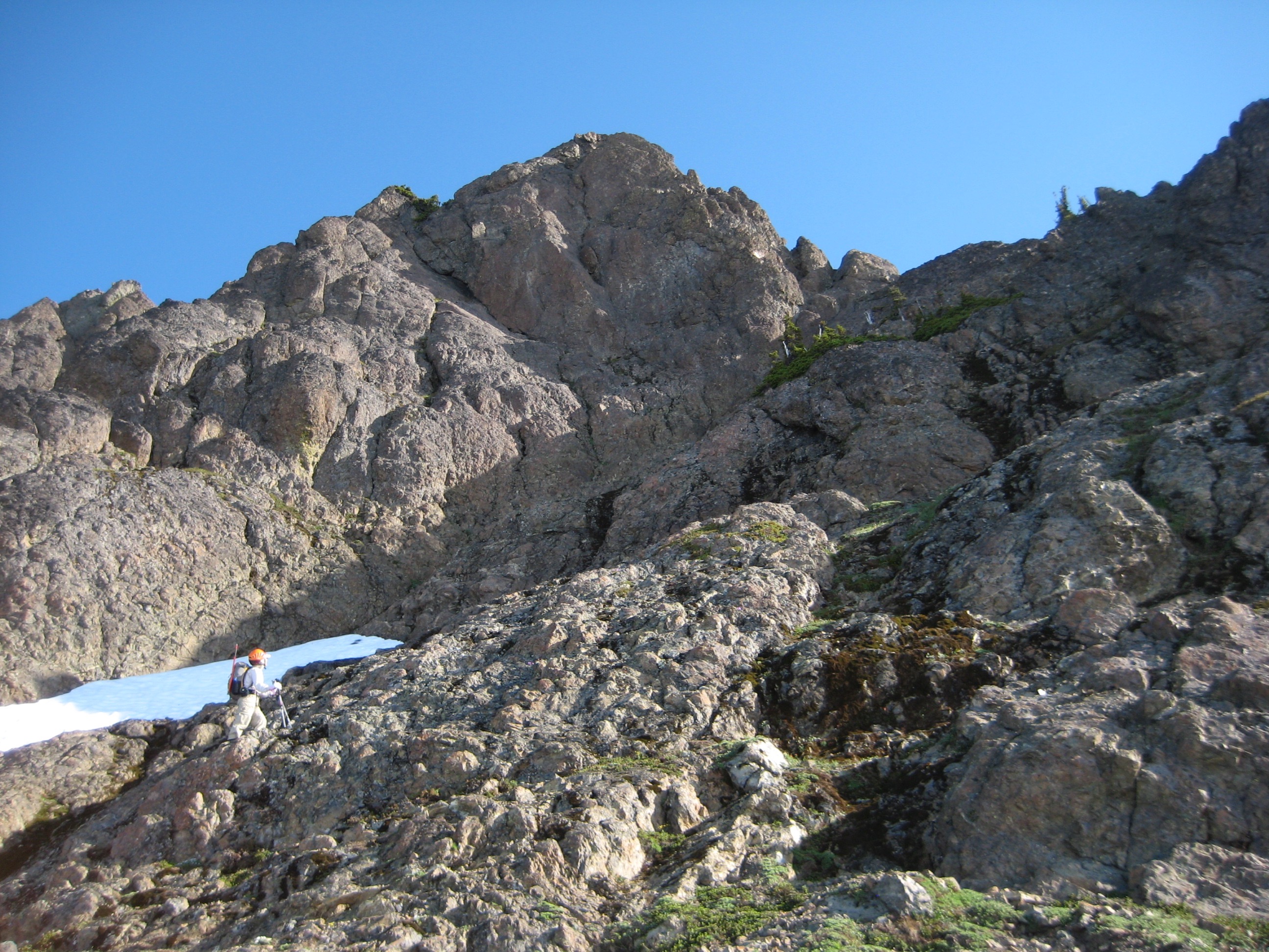 climber scrambling rock toward SE Ridge of North Brother with snow fields in the background