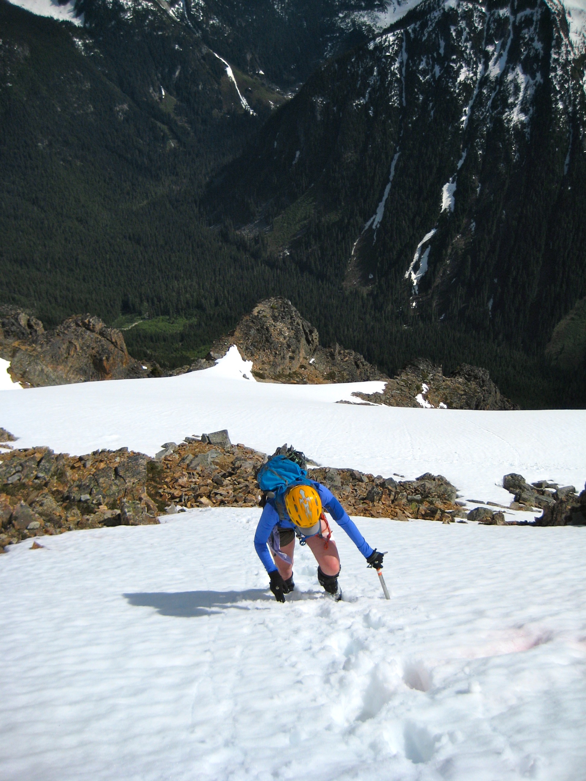 looking down on mountain climber booting up snow slope with forest floor below the summit of Kimtah Peak in the North Cascades