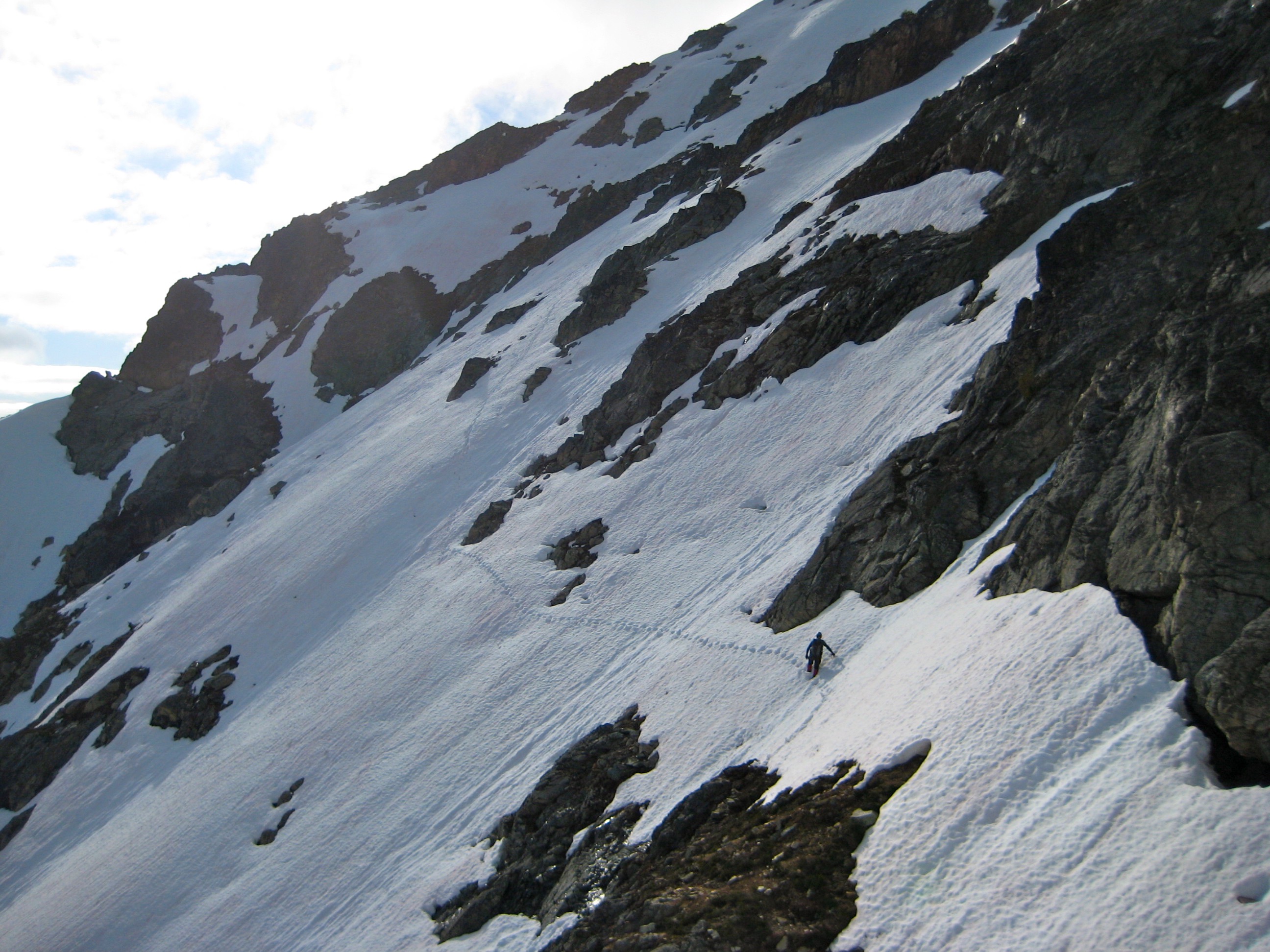 mountain climber traversing steep snow patches with exposed rock slabs on the sholder of Kimtah Peak in the North Cascades