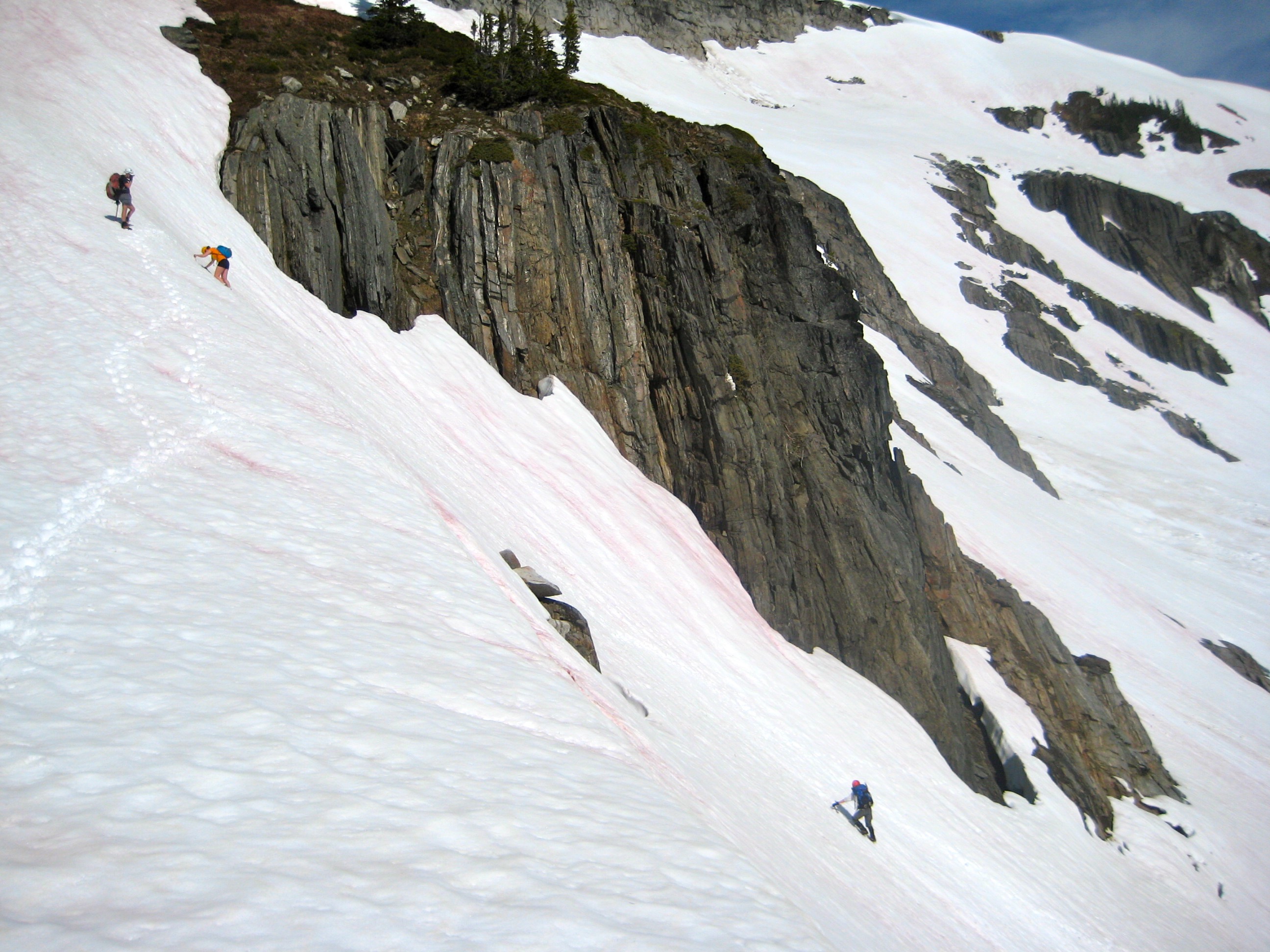 mountain climbers facing in booting down steep snow slope below Boulder Pass in the Glacier Peak Wilderness with a steep rock wall in the background