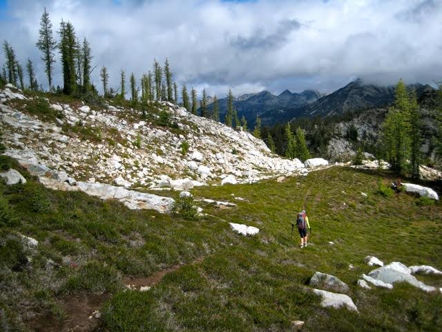 hikers crossing Rainbow Ridge grassy meadow with a rocky boulder shoulder in the Stehekin Mountains