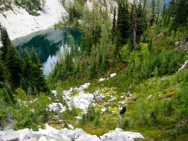 hikers scrambling down boulder field towards a lake in an alpine forest in the Stehekin Mountains