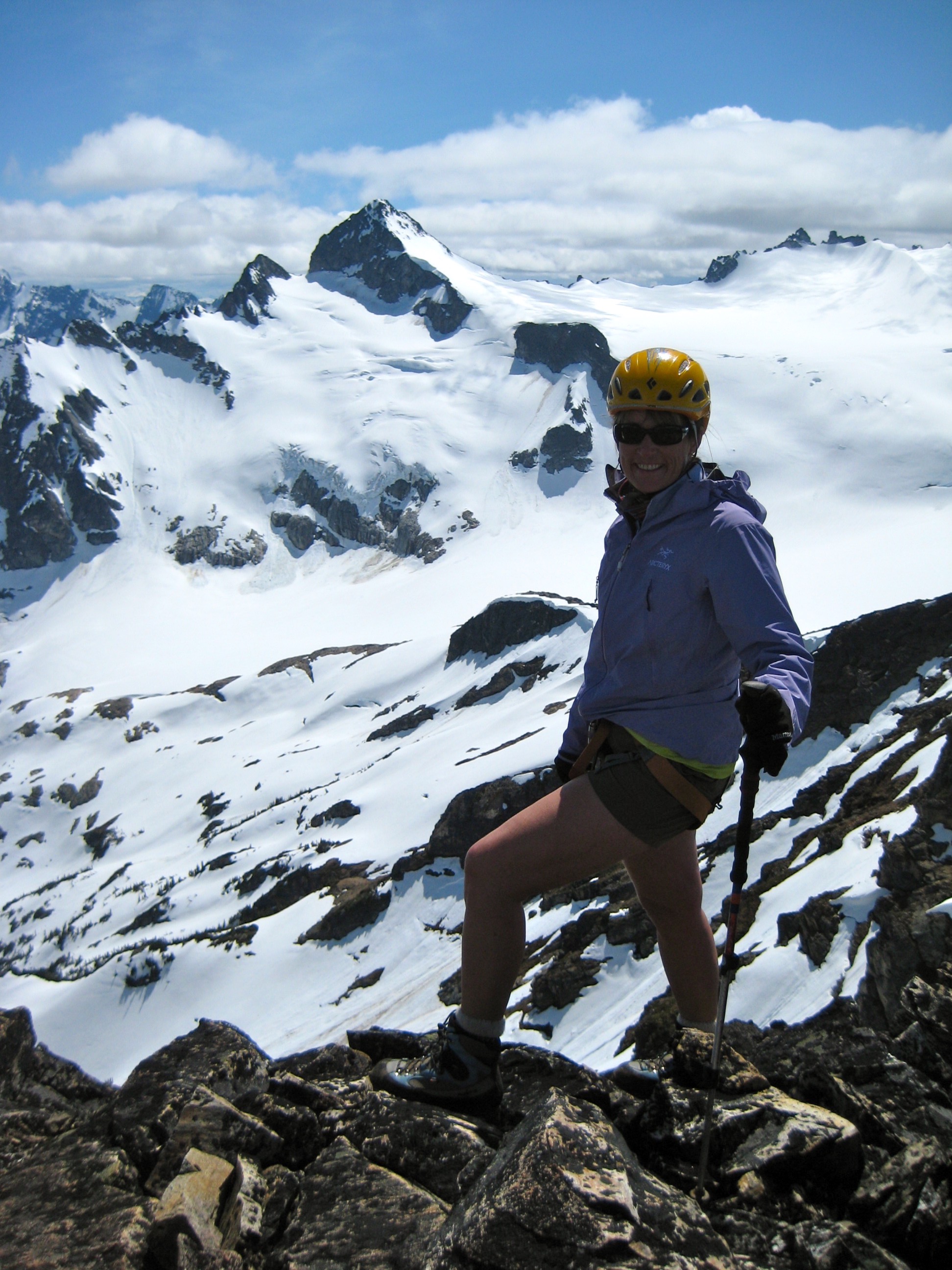 mountain climber stands on the rocky, summit of Colonial Peak with Snowfield Peak in the McAllister Mountains in the background 