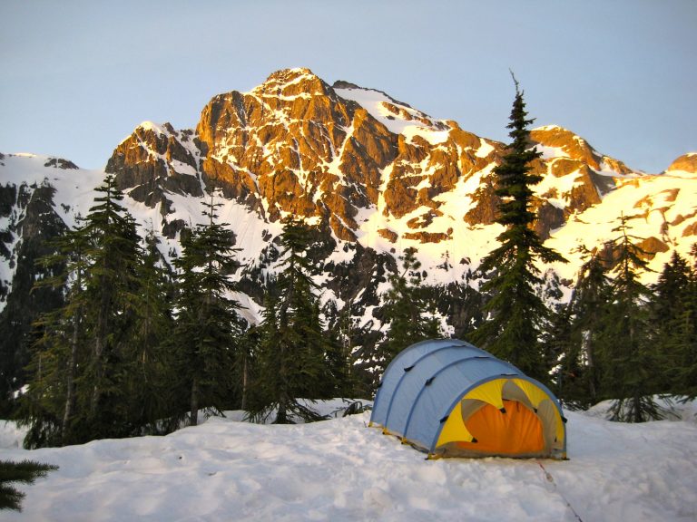 Morning sun warms the fluted face of Colonial Peak in the McAllister Mountains with a mountain climber's tent pitched on a snowy hill in North Cascades National Park