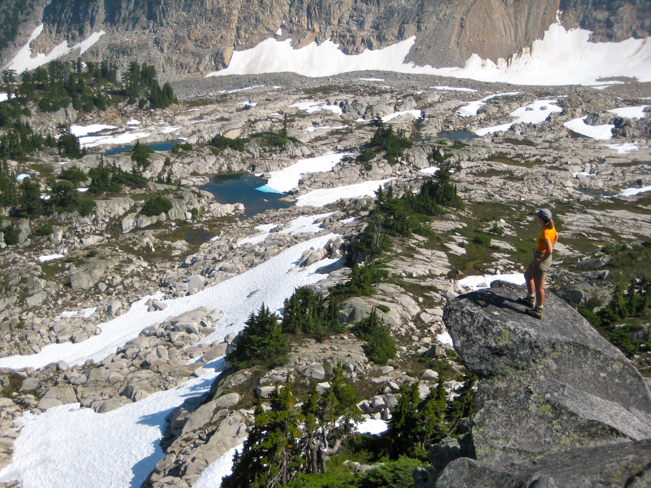 backpacker standing on a rock outcropping looking across rock slabs and linguring snow fields at the Tank Lakes Tarns in the Snoqualmie Mountains
