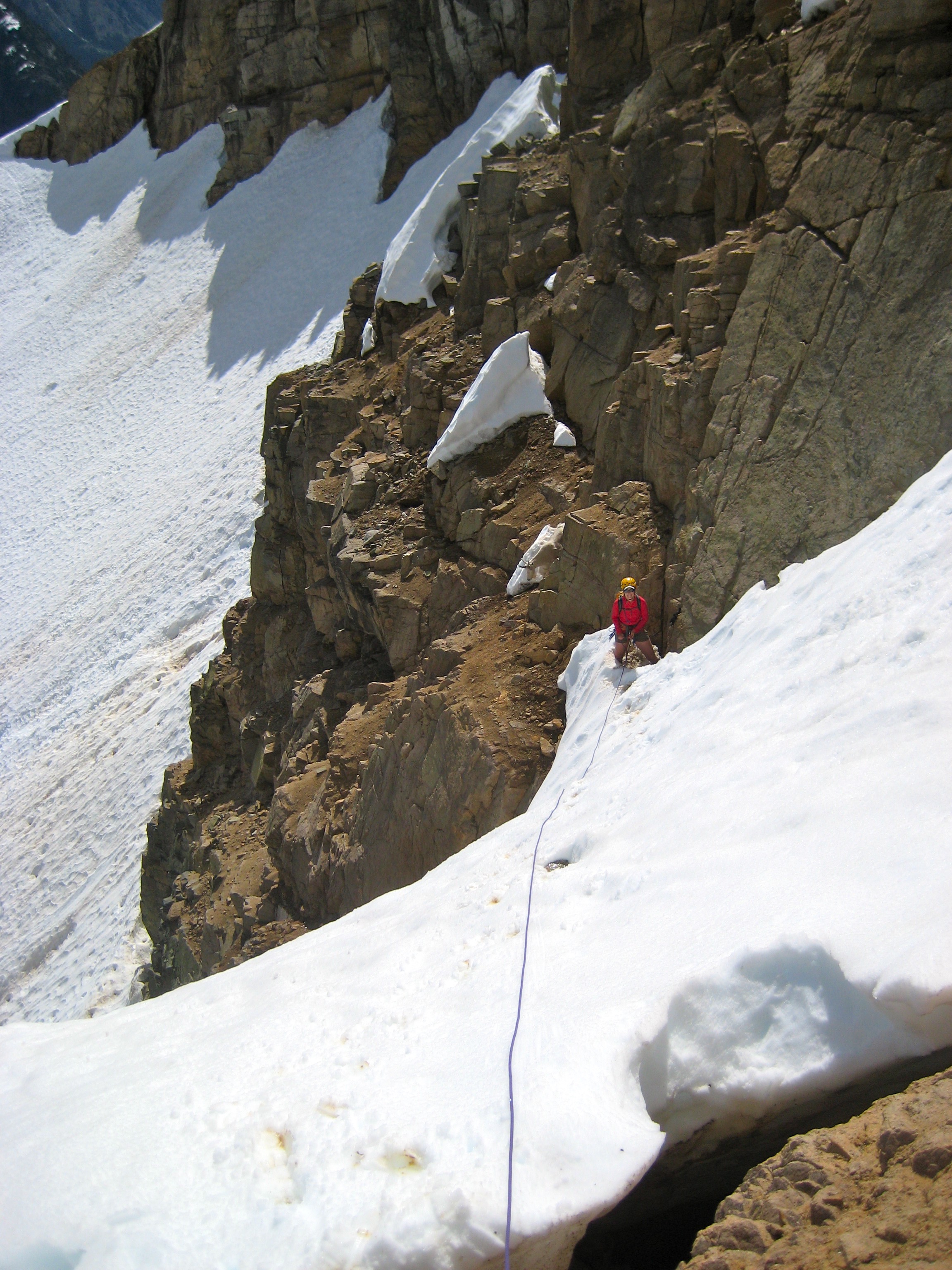 mountain climber belaying from sandy, rock ledges across snow feilds on the shoulder of Corteo Peak in the Ragged Ridge Mountains in North Cascades National Park