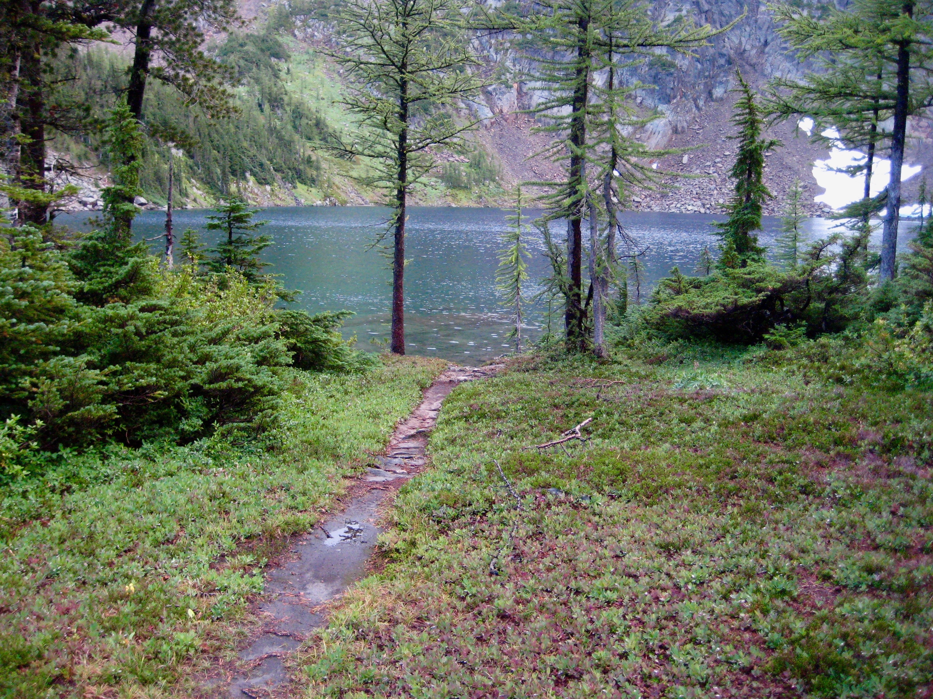 trail leading to Larch Lake in the Chiwaukum Mountains with evergreen trres and a linguring snow patch in the Alpine Lakes WIlderness