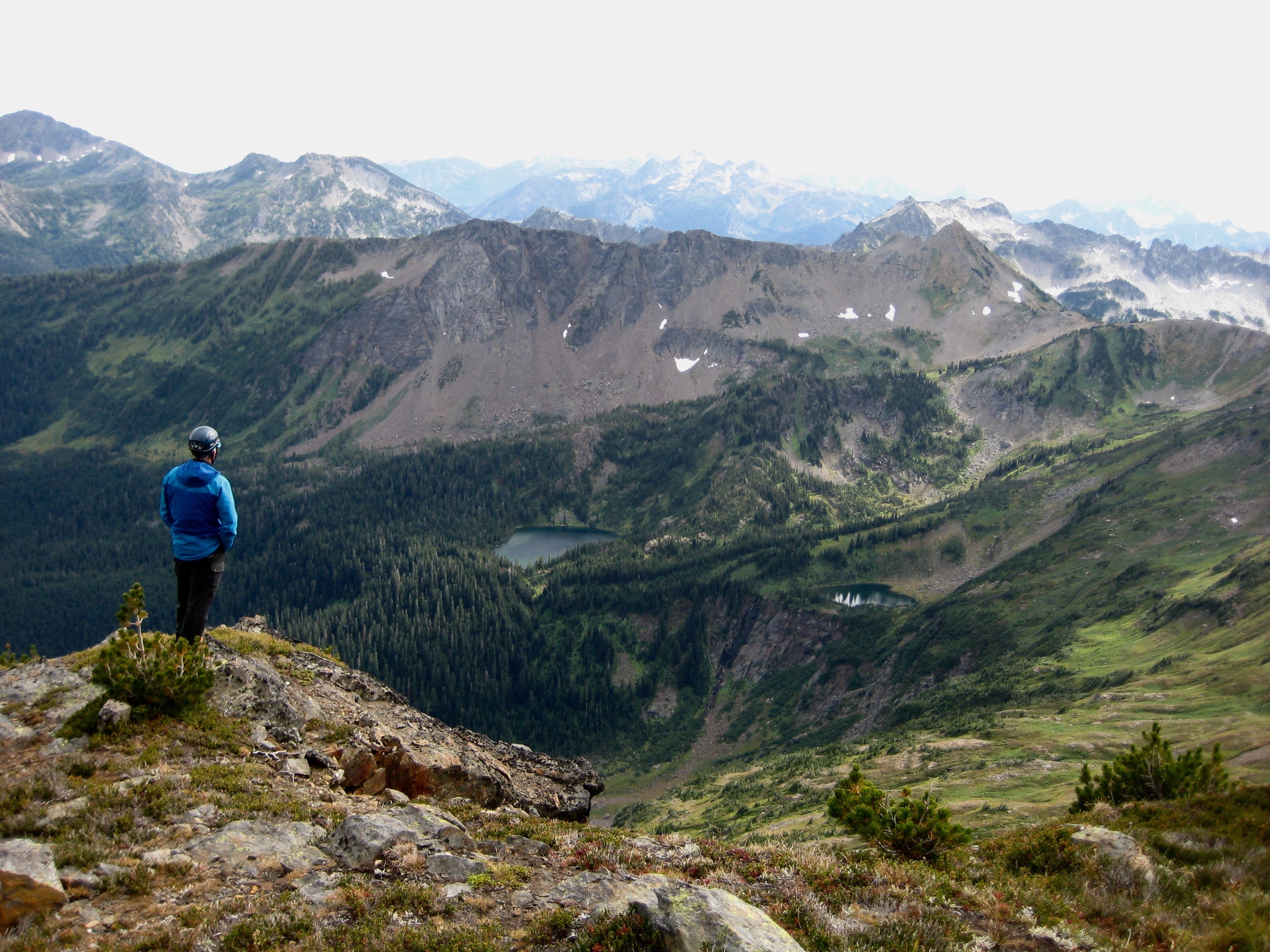 mountain climber standing apon rocky bump looking down on Lake Flora, Lake Brigham, and the Chiwaukum Mountains in the Alpine Lakes Wilderness