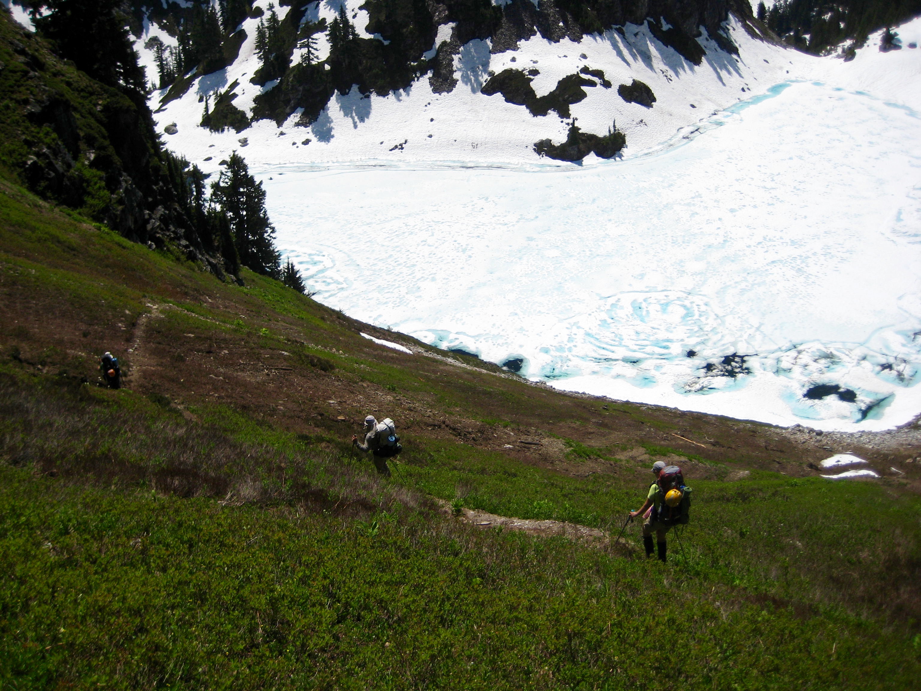 looking down on mountain climbers traversing steep grassy slopes above snow covered cub lake in the Ptarmingan Mountains