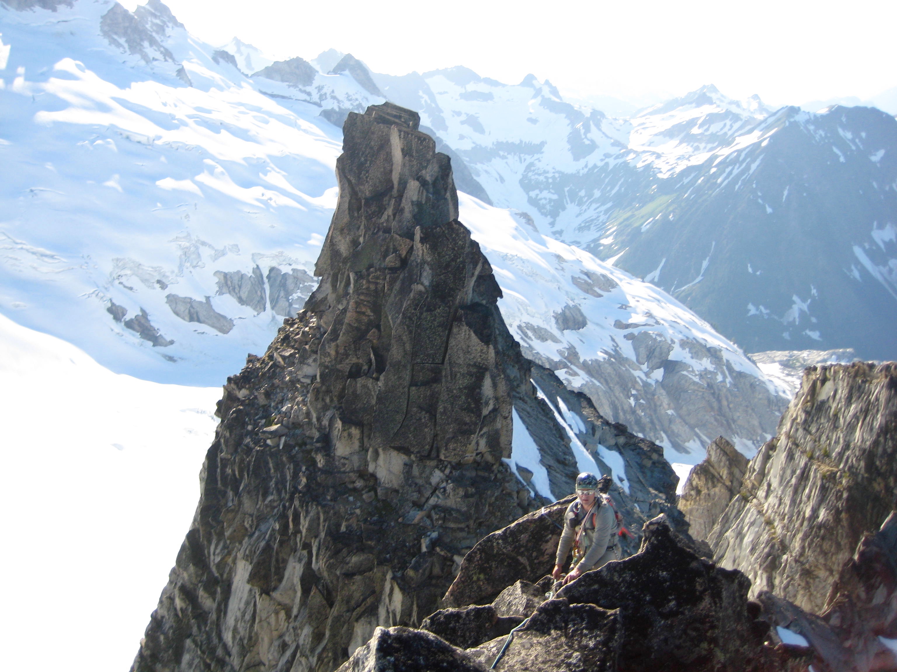roped mountain climber scrambles steep ridge crest with rock horns of Gunsight Peak with the Ptarmigan Mountains in the background