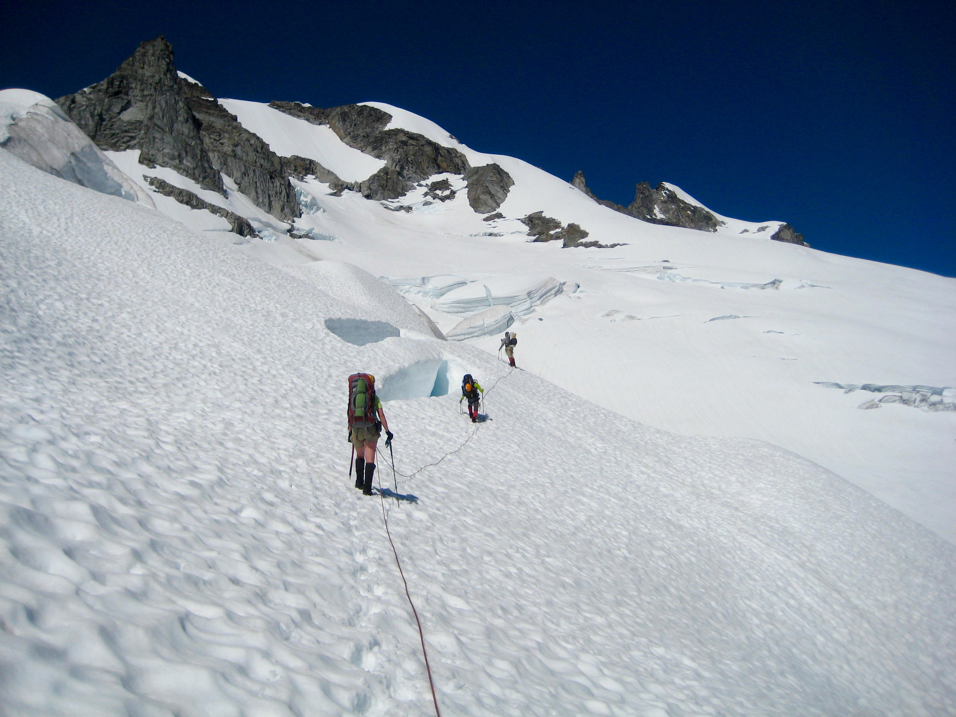 roped mountain climbers traverse snow slope with minor cravasses under Sinister Peak in the Ptarmigan Mountains