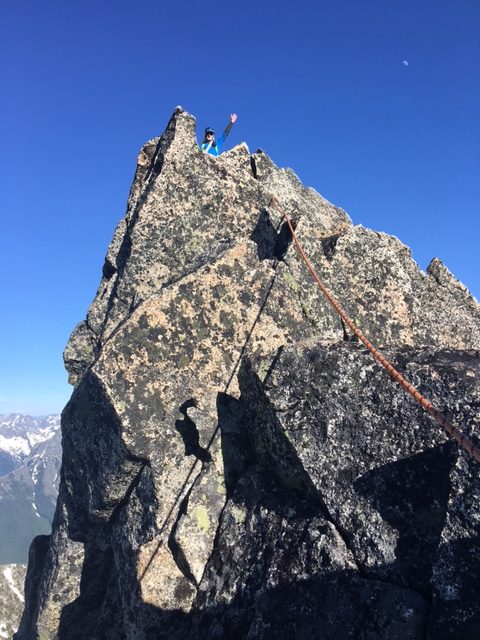looking up at roped, waving mountain climber on the rock horn summit of Gunsight Peak in the Glacier Peak WIlderness 